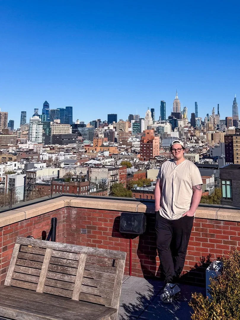 A person with glasses and tattoos, wearing a light shirt and black pants, standing on a rooftop with a view of New York City skyline in the background, including the Empire State Building, under a clear blue sky.
