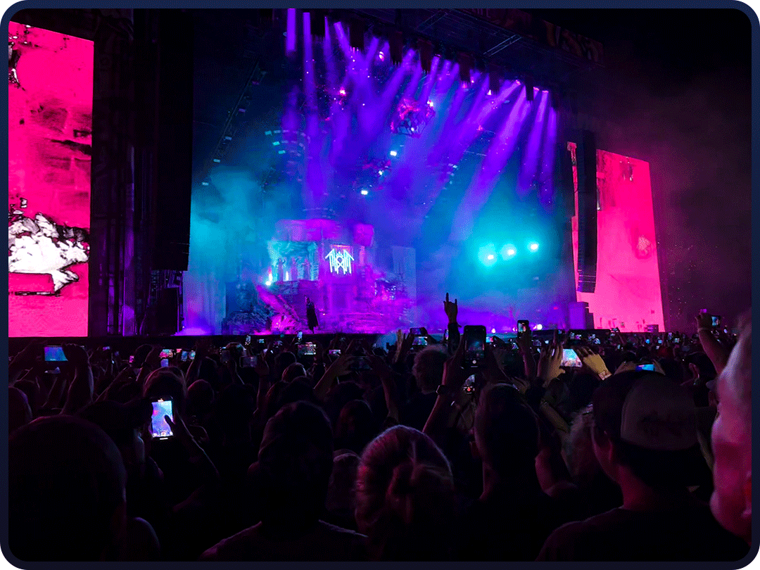Crowd at a concert with colorful stage lights, large screens, and audience members taking photos.