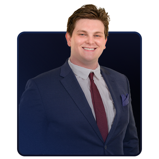 A young man in a navy blue suit, white shirt, and red tie, smiling and standing against a dark background.