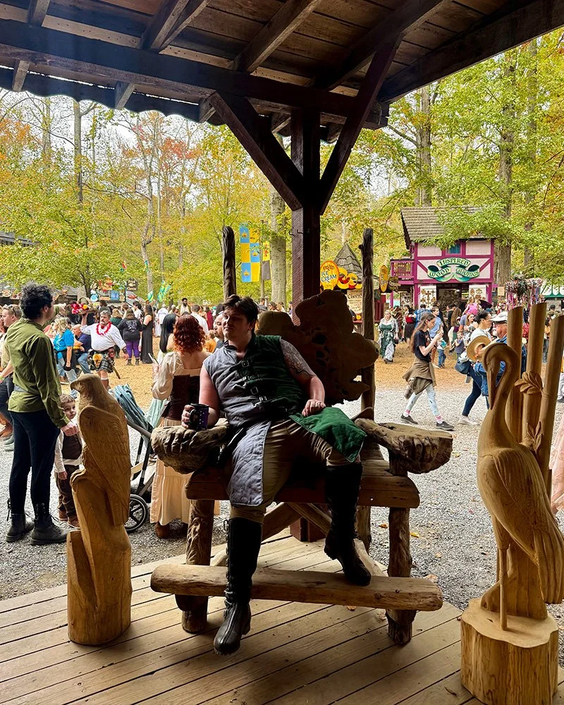 Person sitting on a rustic wooden bench under a wooden shelter at a fair or festival, with trees and people in the background.