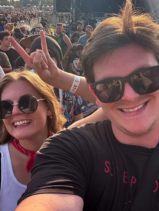 Two people smiling at a crowded outdoor music festival, one making a peace sign behind the other's head.