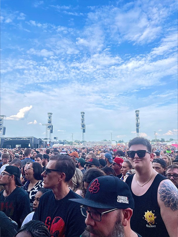 Crowd of people attending an outdoor concert or festival under a partly cloudy sky with four large stage speakers in the background.