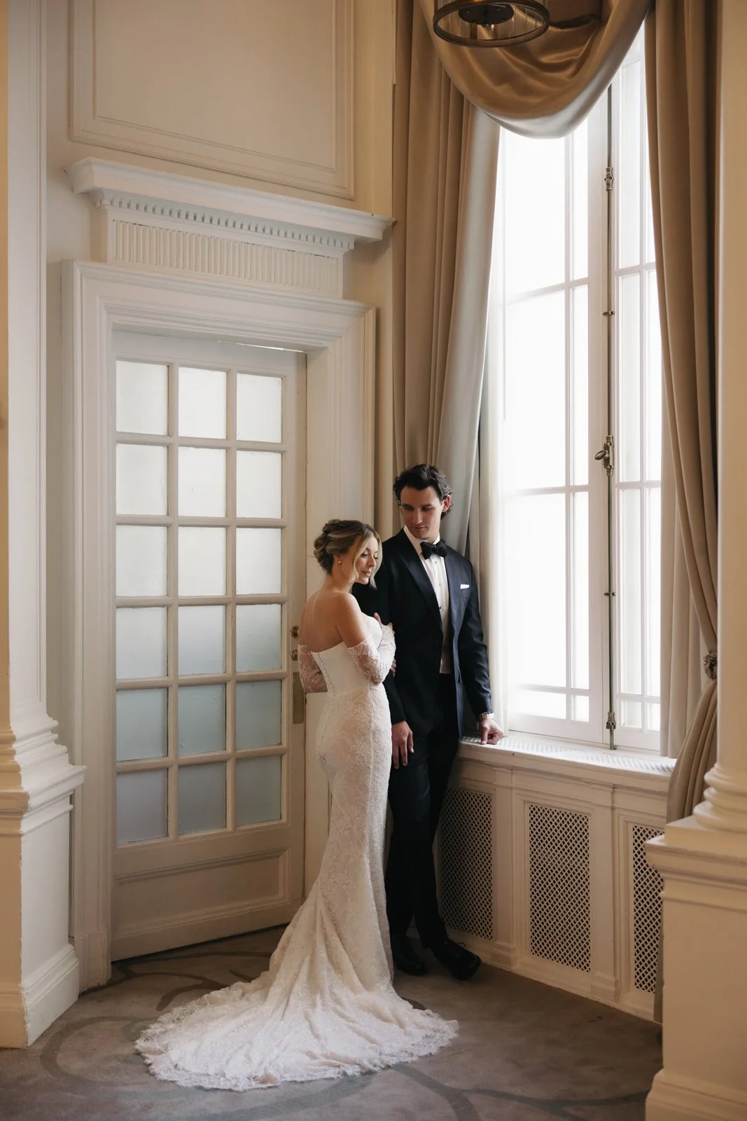 Bride and groom in wedding attire standing by large window with curtains in an elegant room.