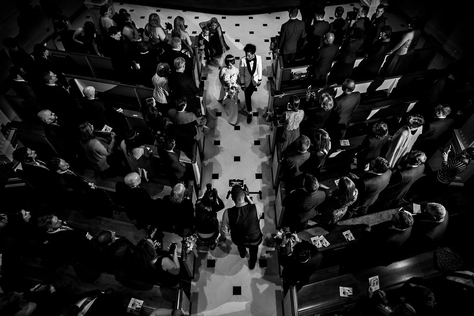Black and white photo of a wedding ceremony inside a church, with a bride and groom walking down the aisle surrounded by seated guests.