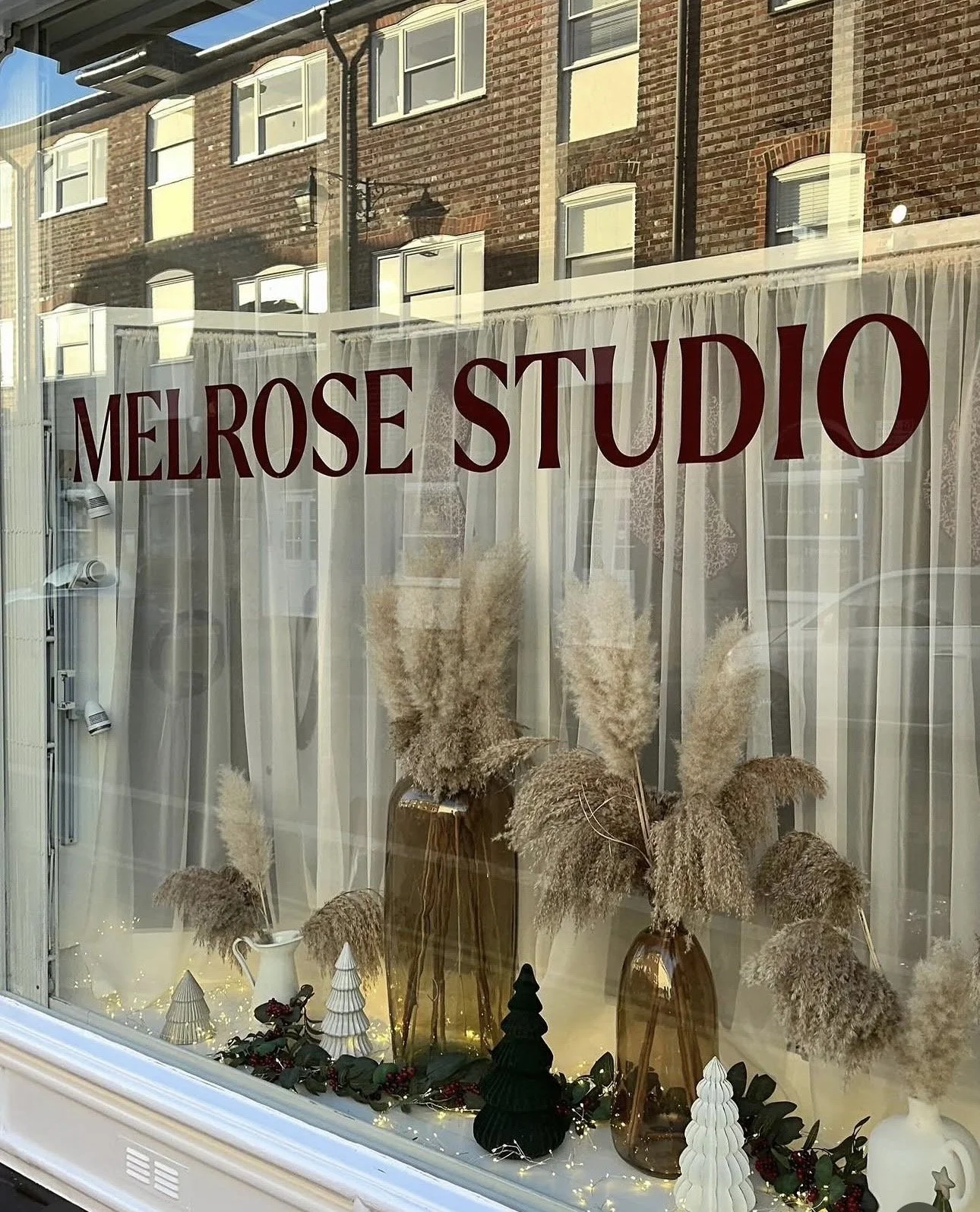 Display window with white curtains, decorative plants in brown vases, and holiday ornaments, with a sign reading 'Melrose Studio' in red capital letters.