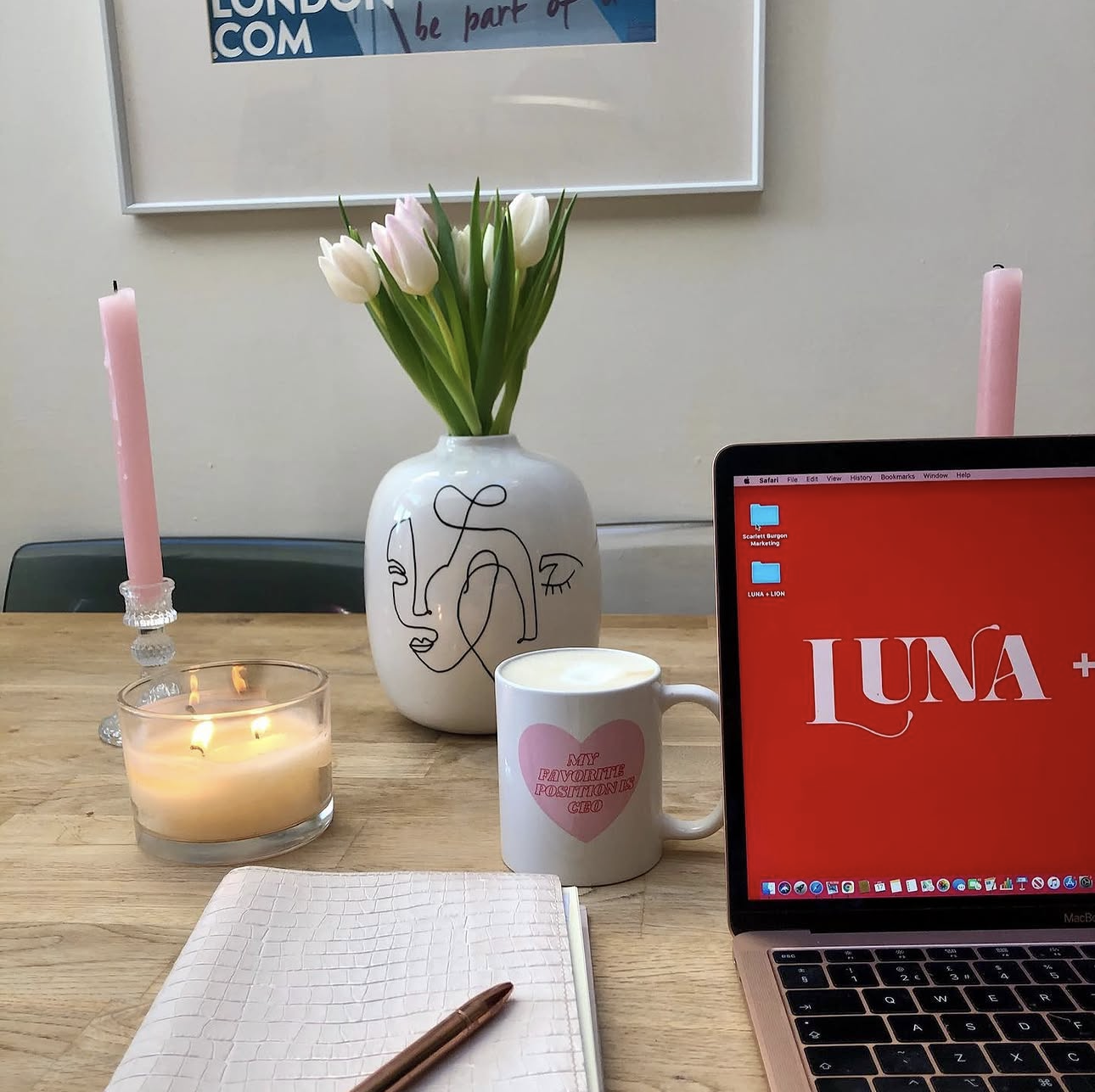 A wooden desk with a laptop displaying the word "LUNA+" on a red background, a white mug with a pink heart and the text "My favorite florals in CBD," a lit candle, a notepad with a pen, a white vase with line art face design holding pink and white tulips, and two pink candles in glass holders. A framed poster is on the wall in the background.
