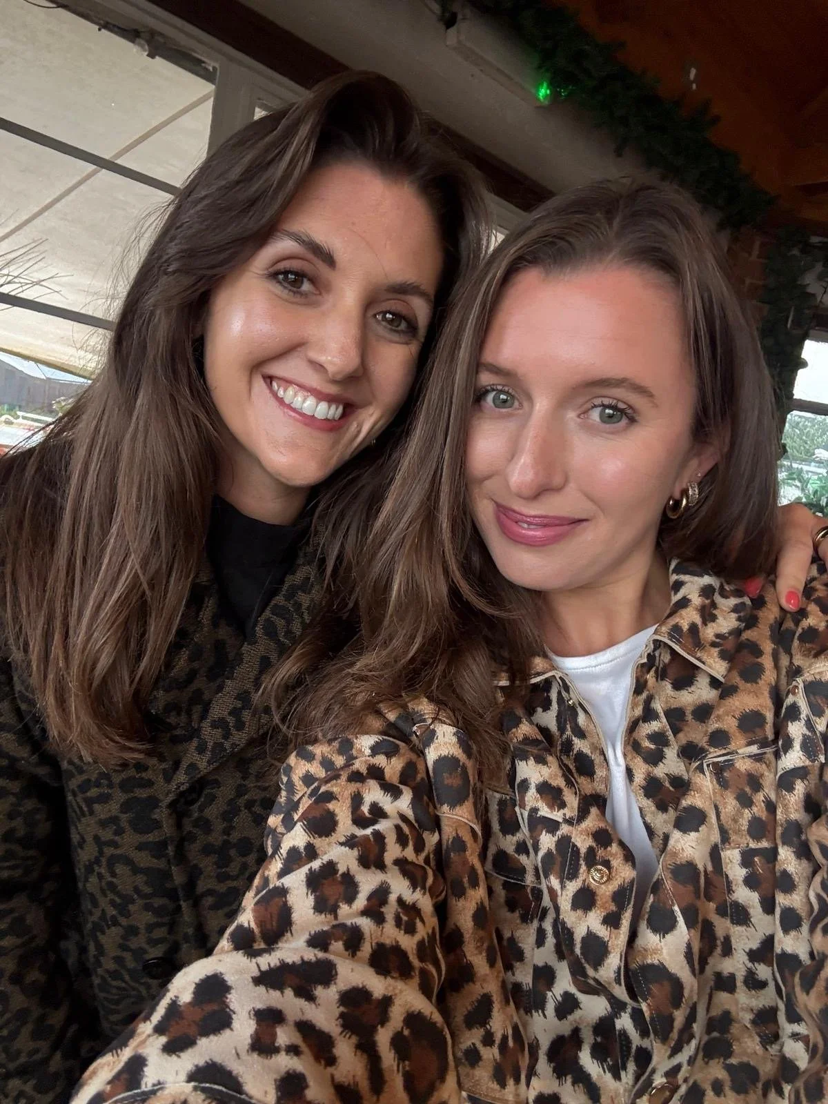 Two women with long brown hair, wearing leopard print jackets, smiling and posing for a selfie indoors with wooden ceiling and windows in the background.