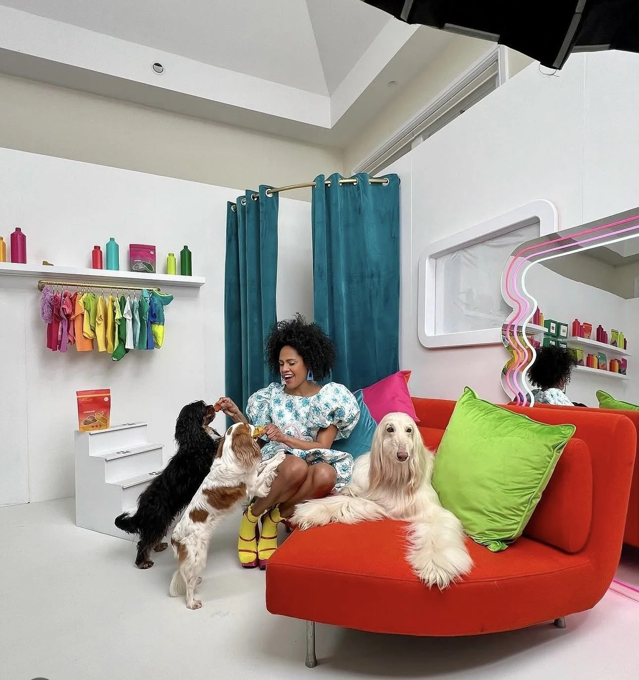 A woman with curly hair sitting on a bright red sofa in a colorful room, playing with three dogs. The room has teal curtains, a wall with rainbow-colored shirts hanging, colorful bottles on shelves, and a mirror with a wavy pink frame.