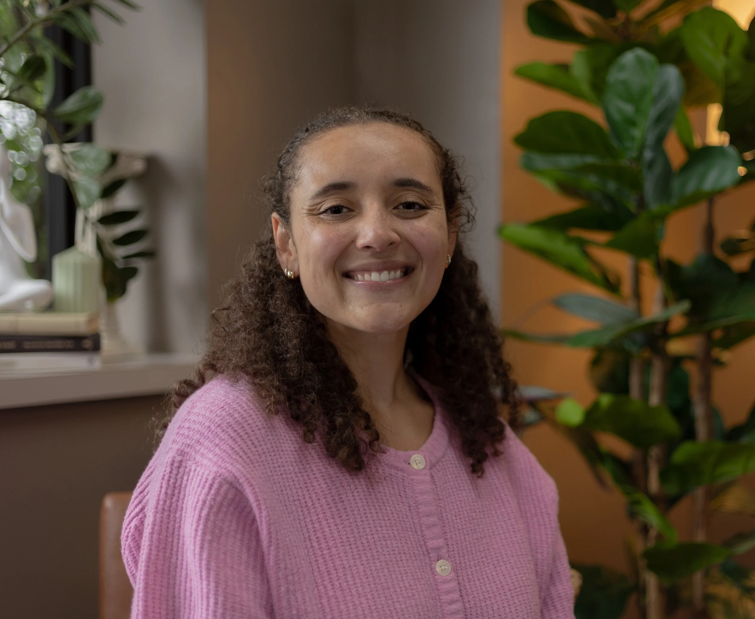 A woman smiling with curly hair, wearing a black top and hoop earrings, standing against a plain gray wall.