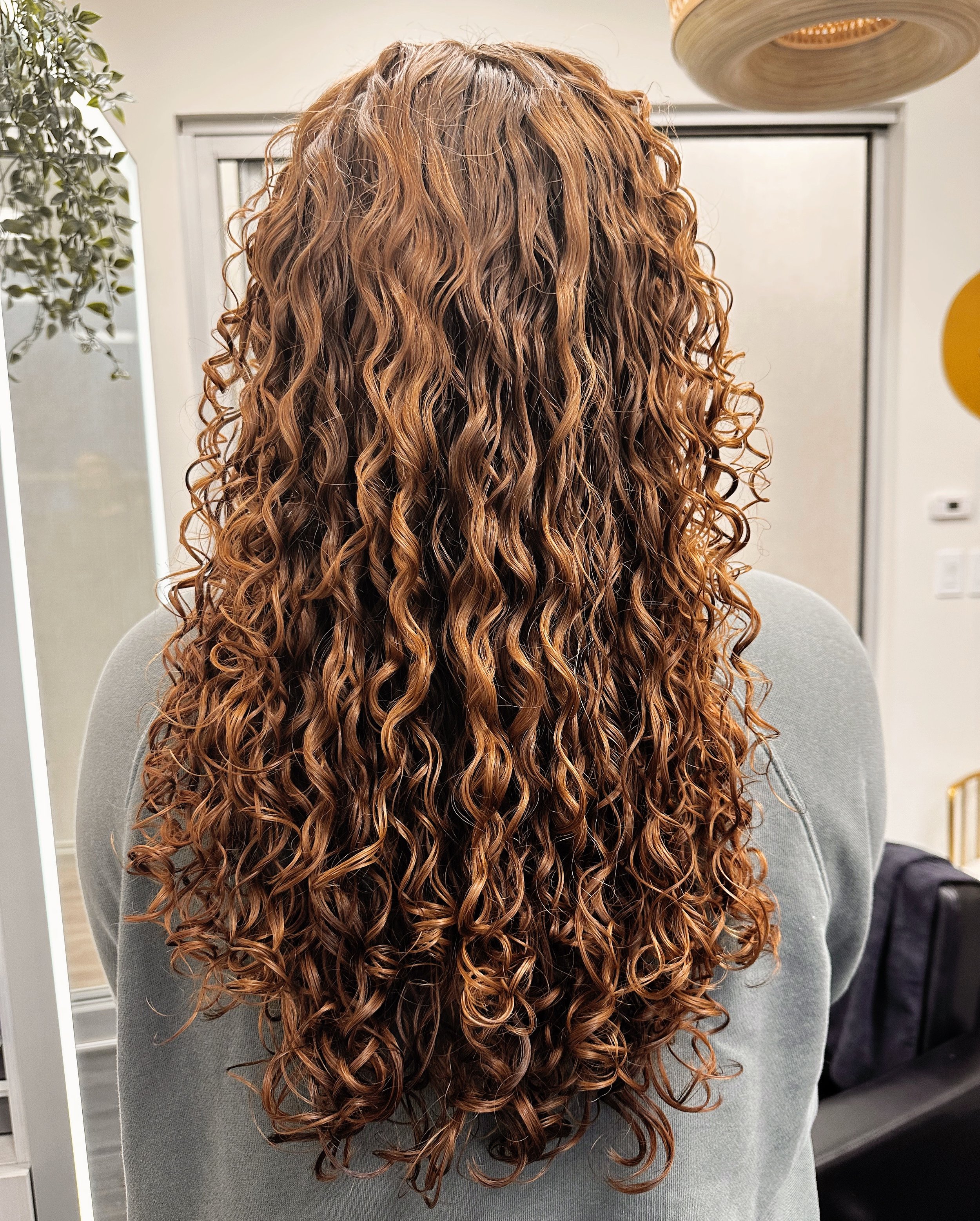 A photo of a client with long curly hair featuring copper-red colors with lots of dimension. 