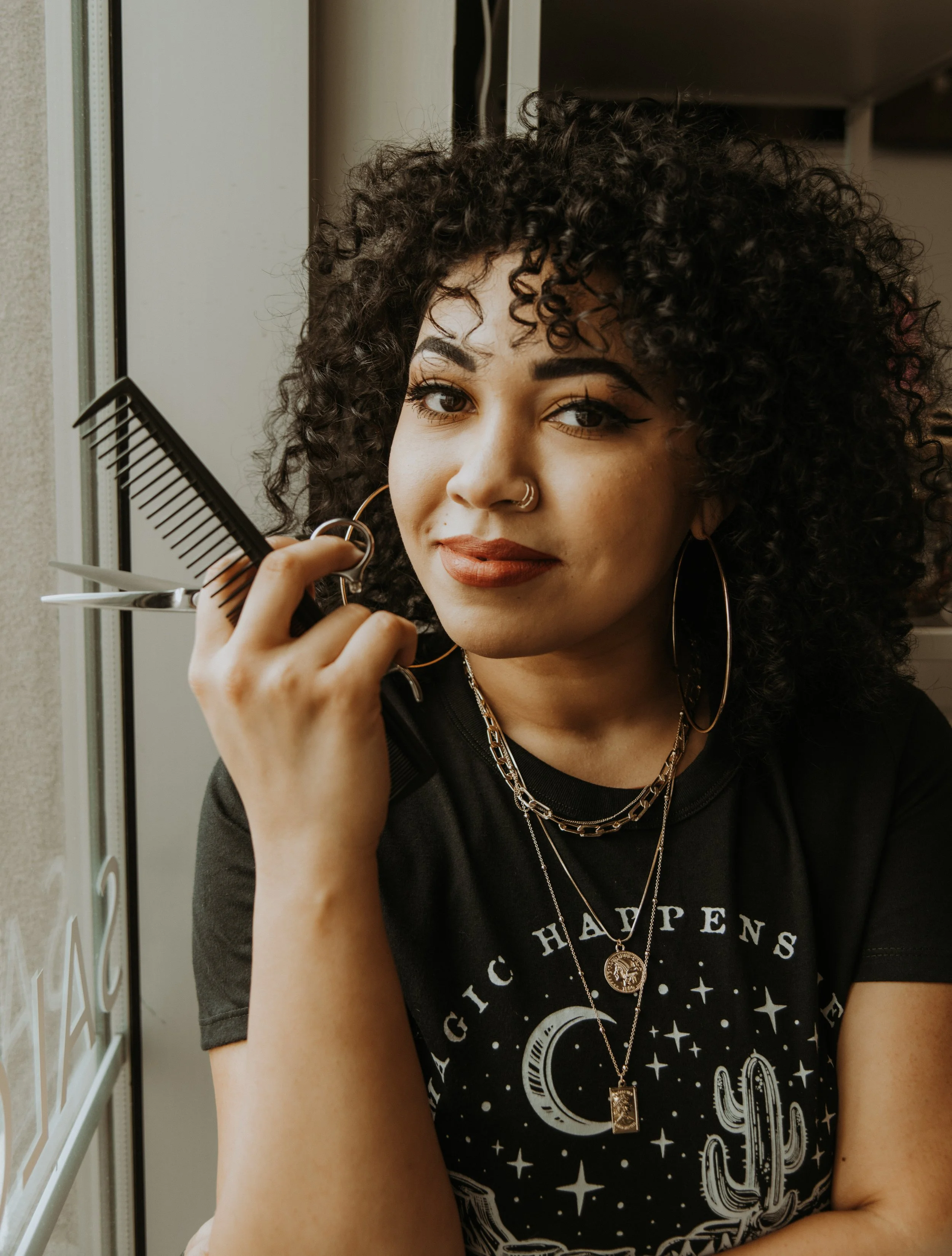 A photo of Ya Girl With the Curl smiling at the camera while holding cutting shears and a black comb.