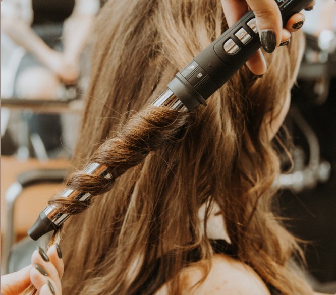 Photo of a stylist using a curling iron on a client with long reddish-brown hair. 