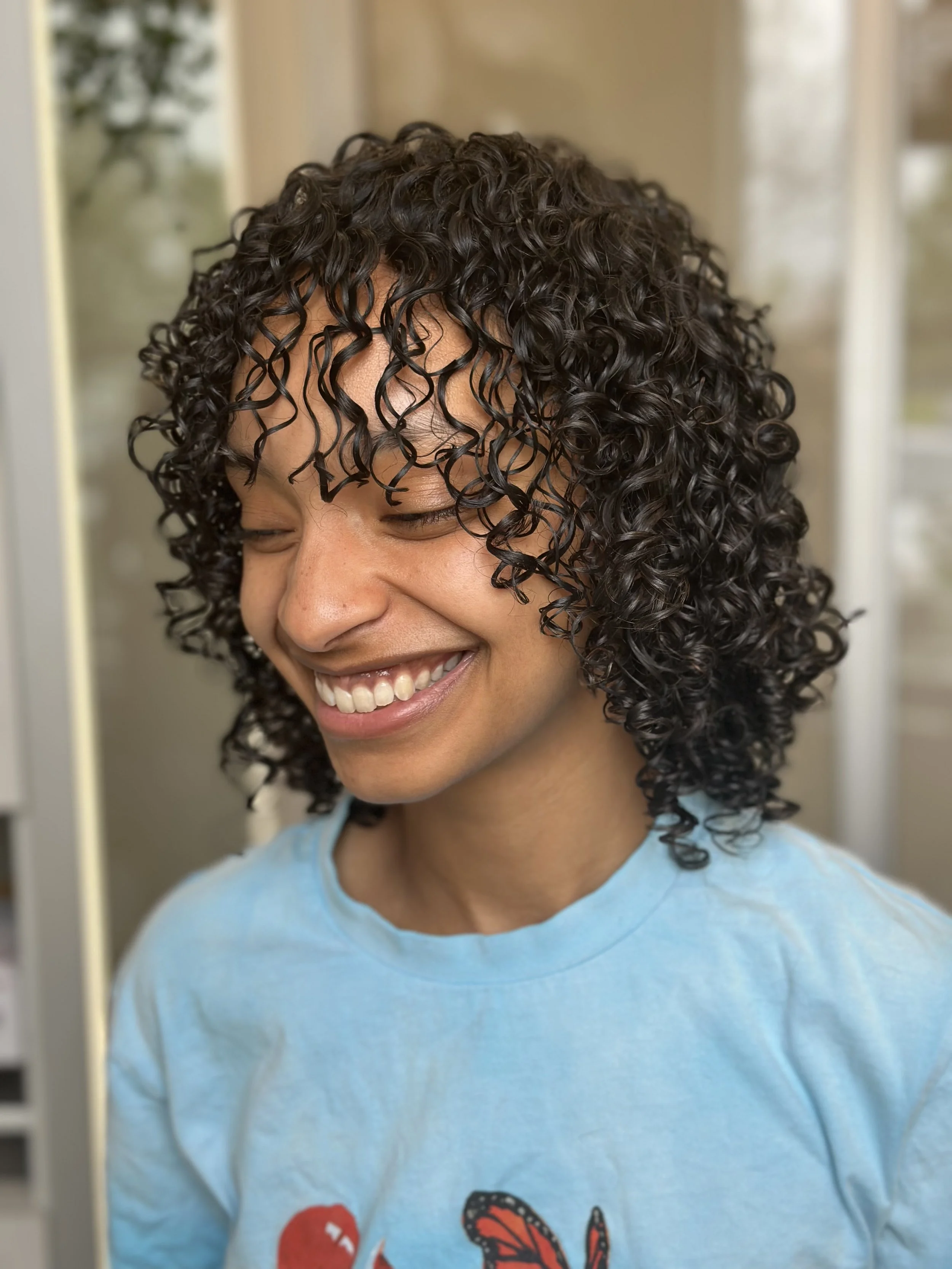 Photo of a smiling client with dark curly shoulder-length hair, including curly bangs and face-framing layers.