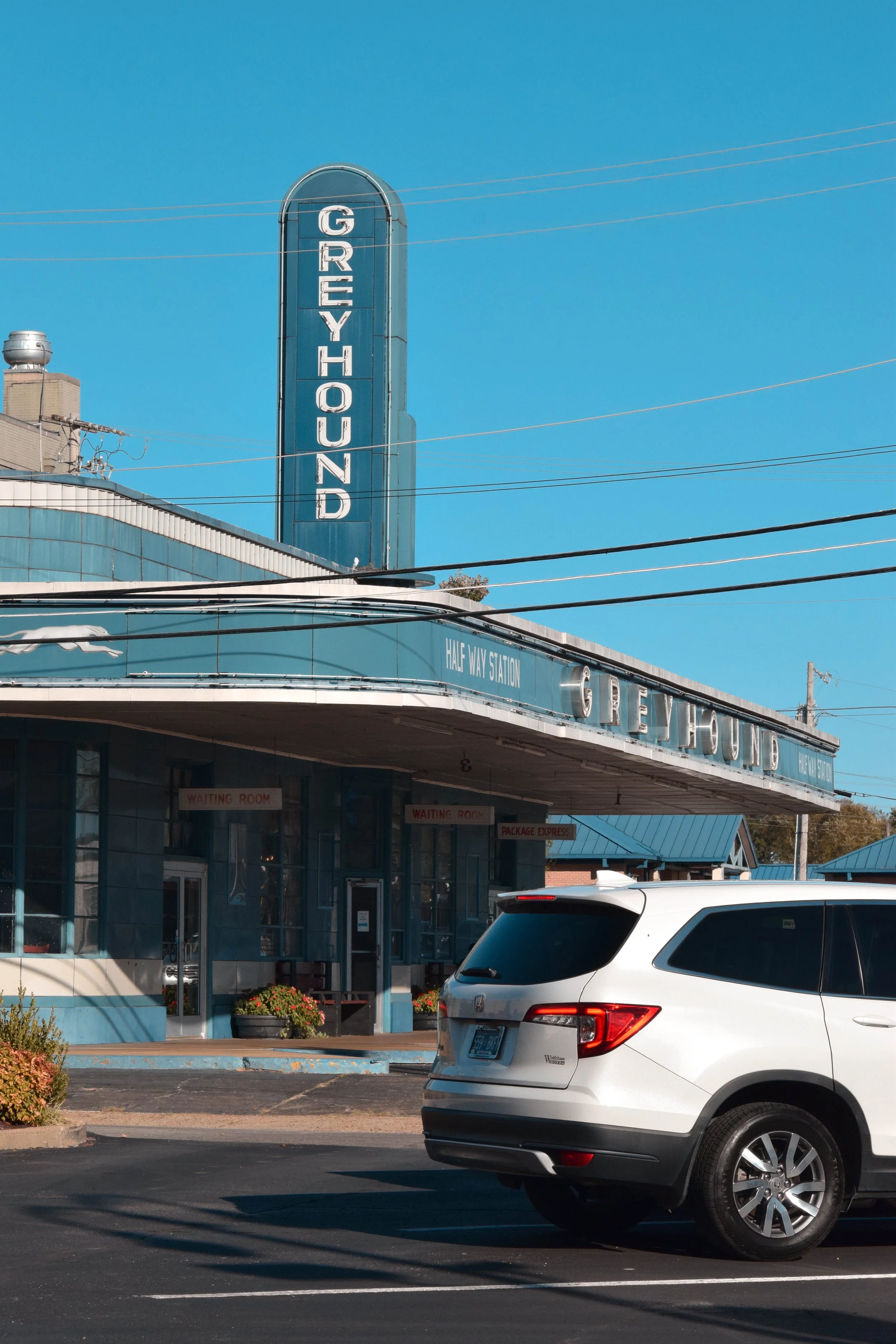 Vintage Greyhound bus station with a large vertical sign reading 'GREYHOUND', a marquee sign with 'HALF WAX STATION', a white SUV parked in front, and a clear blue sky.