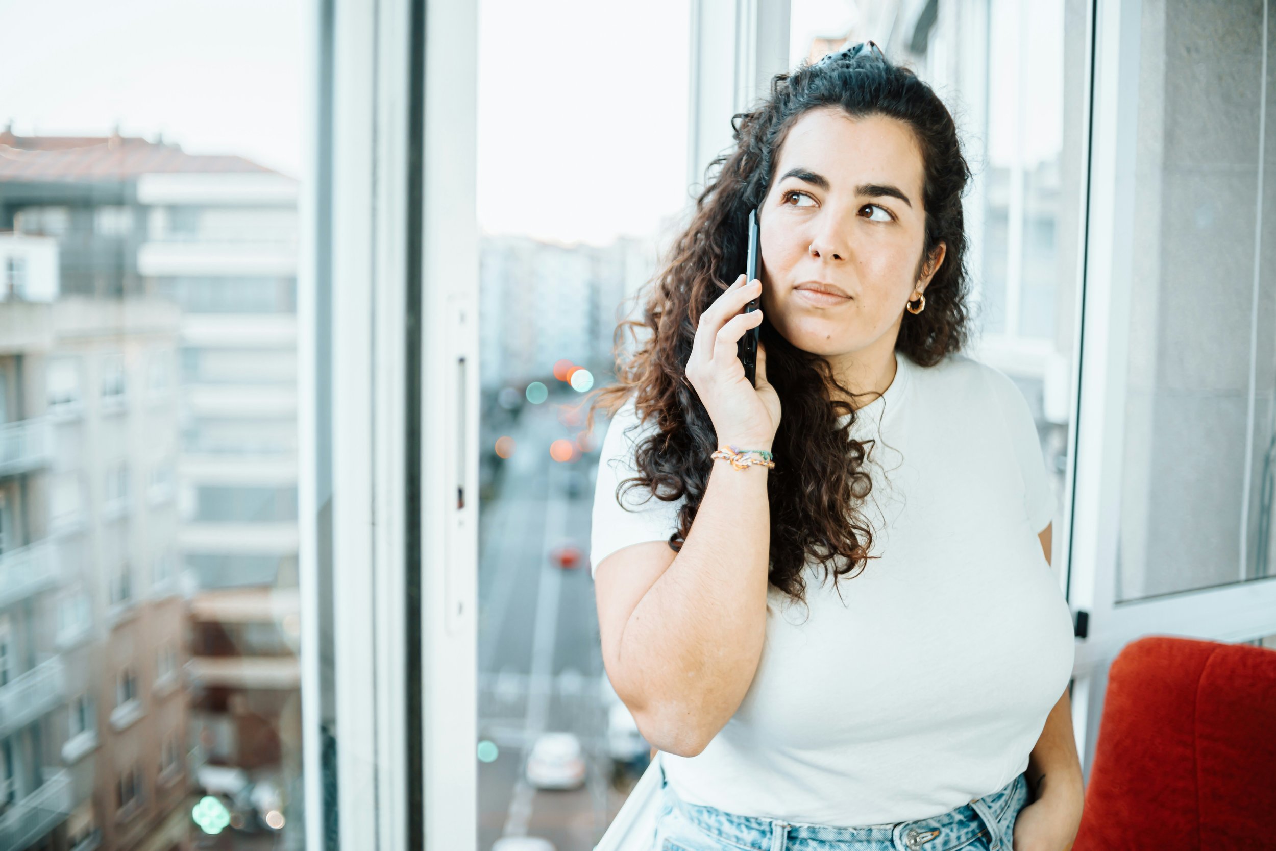 Woman on a phone call by a window looking uncertain, debating between people pleasing or self-loyalty