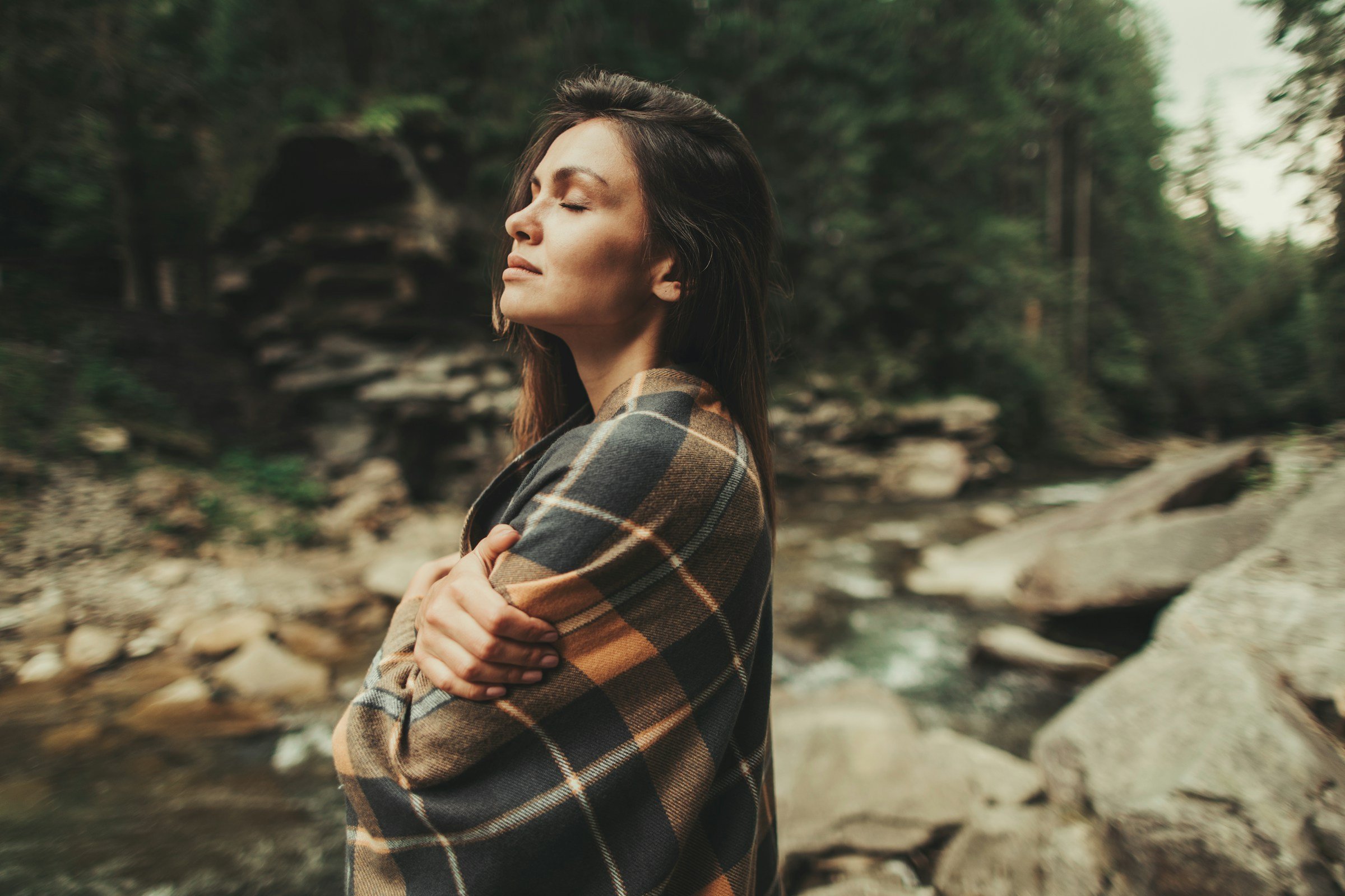 Woman wrapped in plaid blanket in forest looking inward with peaceful expression