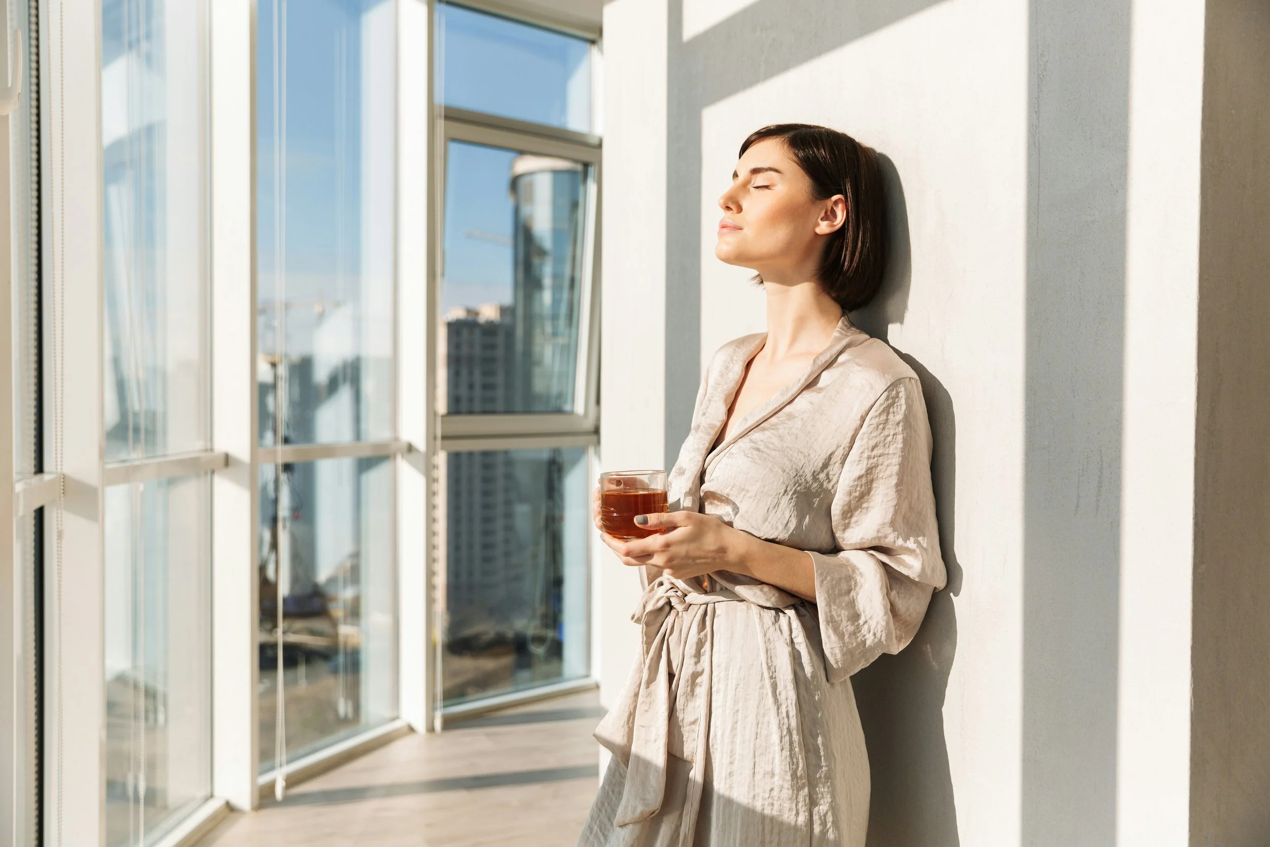 Woman in neutral linen shirt by bright window holding warm beverage