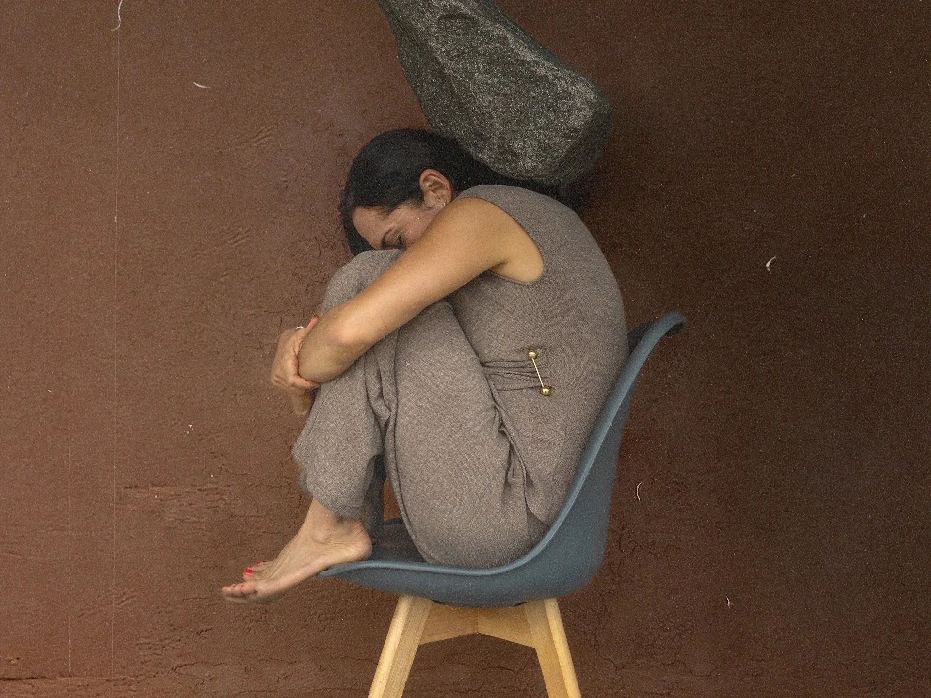 Woman sitting curled inward on a chair against a brown wall