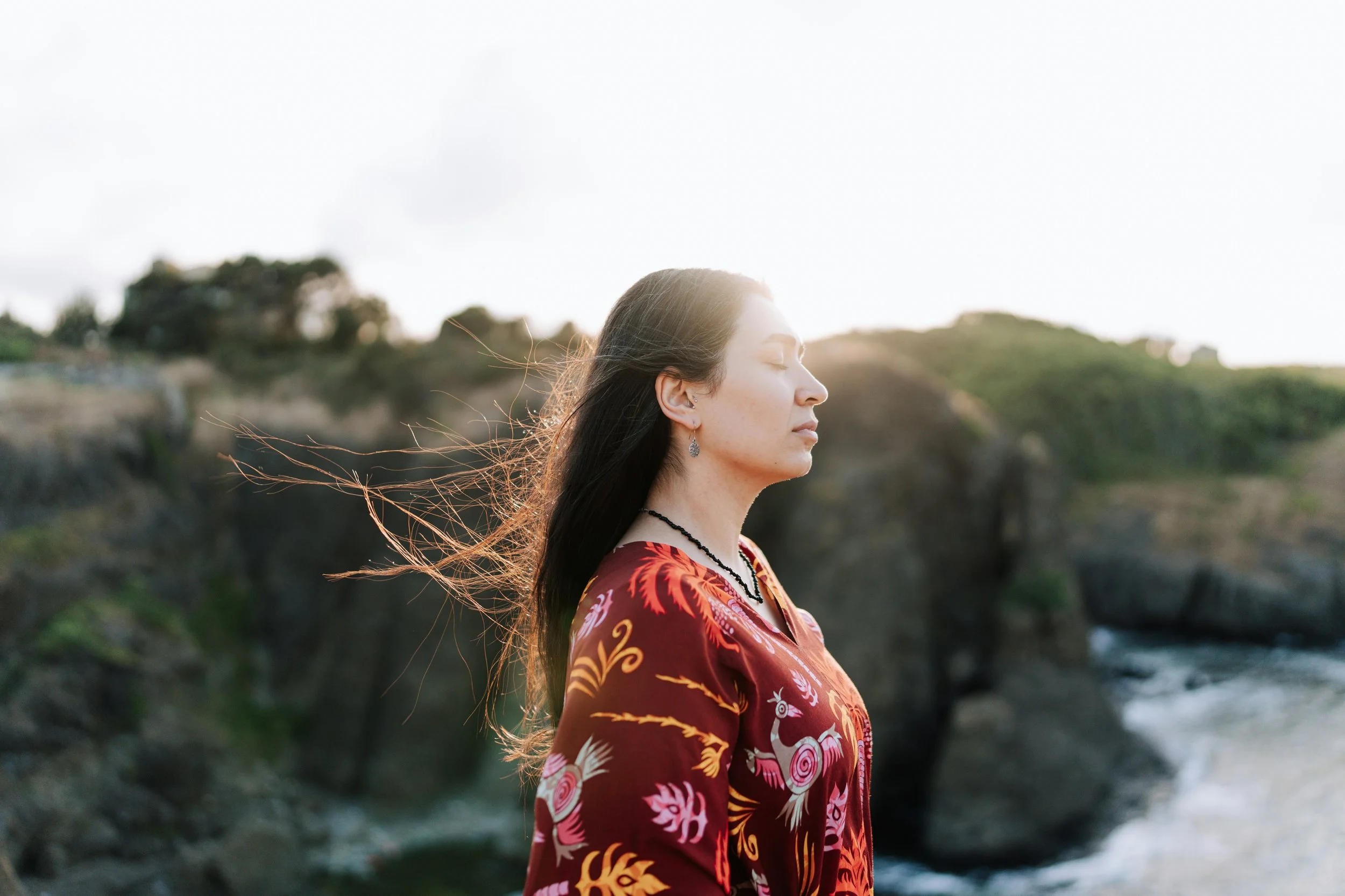 Woman in a red floral dress standing still in wind by a rocky coast, embodying nervous system awareness