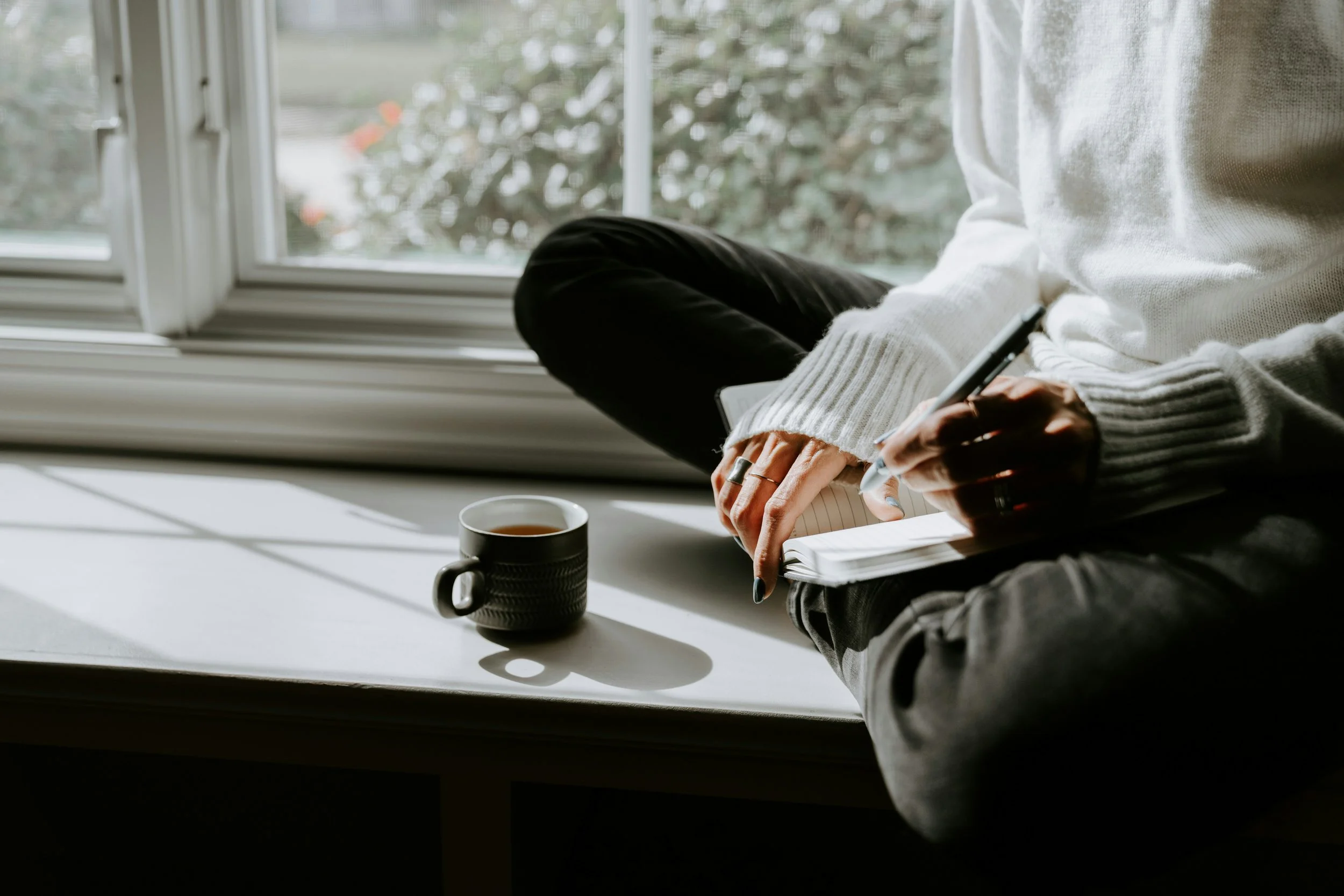 Person sitting cross-legged in a window with a journal and coffee, writing freely in a quiet moment of self-reflection and reconnection.