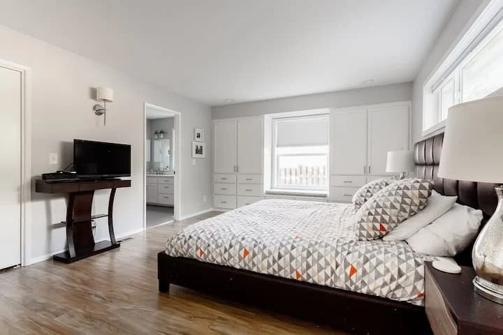 A bedroom with a large bed, geometric pattern bedding, nightstand with a lamp, and large built-in storage cabinets, with a window letting in natural light.