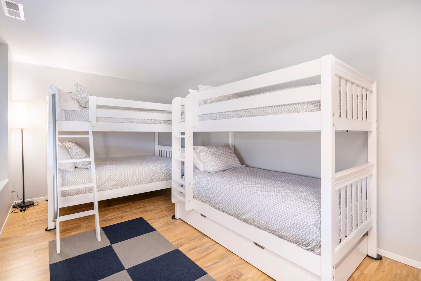 Two white wooden bunk beds with gray and white geometric bedding in a room with a hardwood floor, a small black and gray rug, and a white wall.