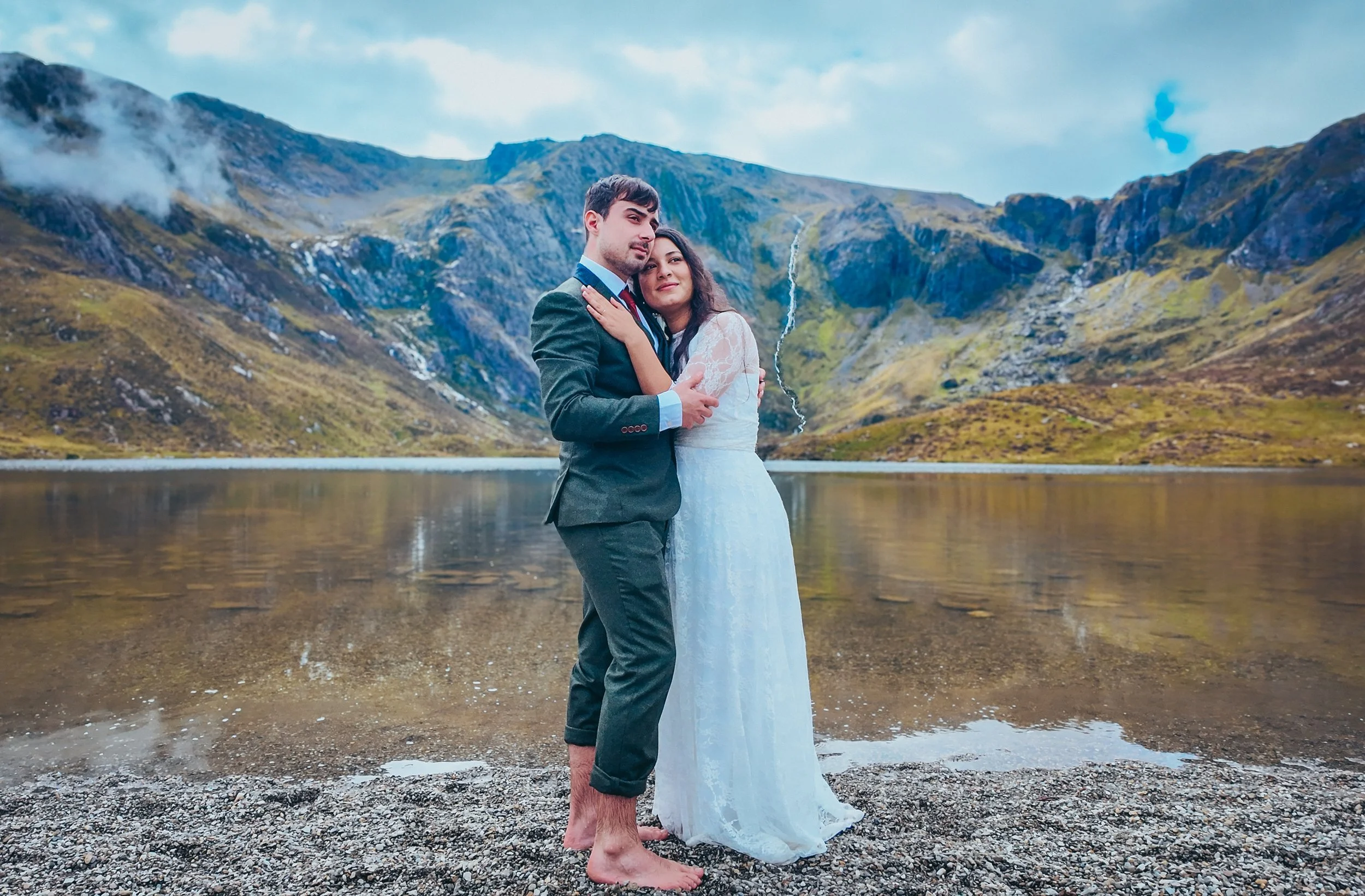 Davinia and Joao embracing barefoot on the shore of a mountain lake