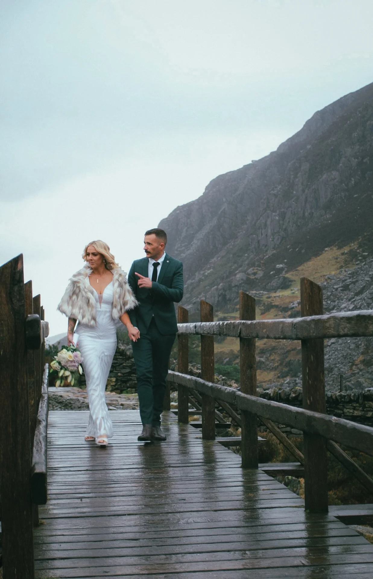 A couple walking along a wooden boardwalk beneath a mountain