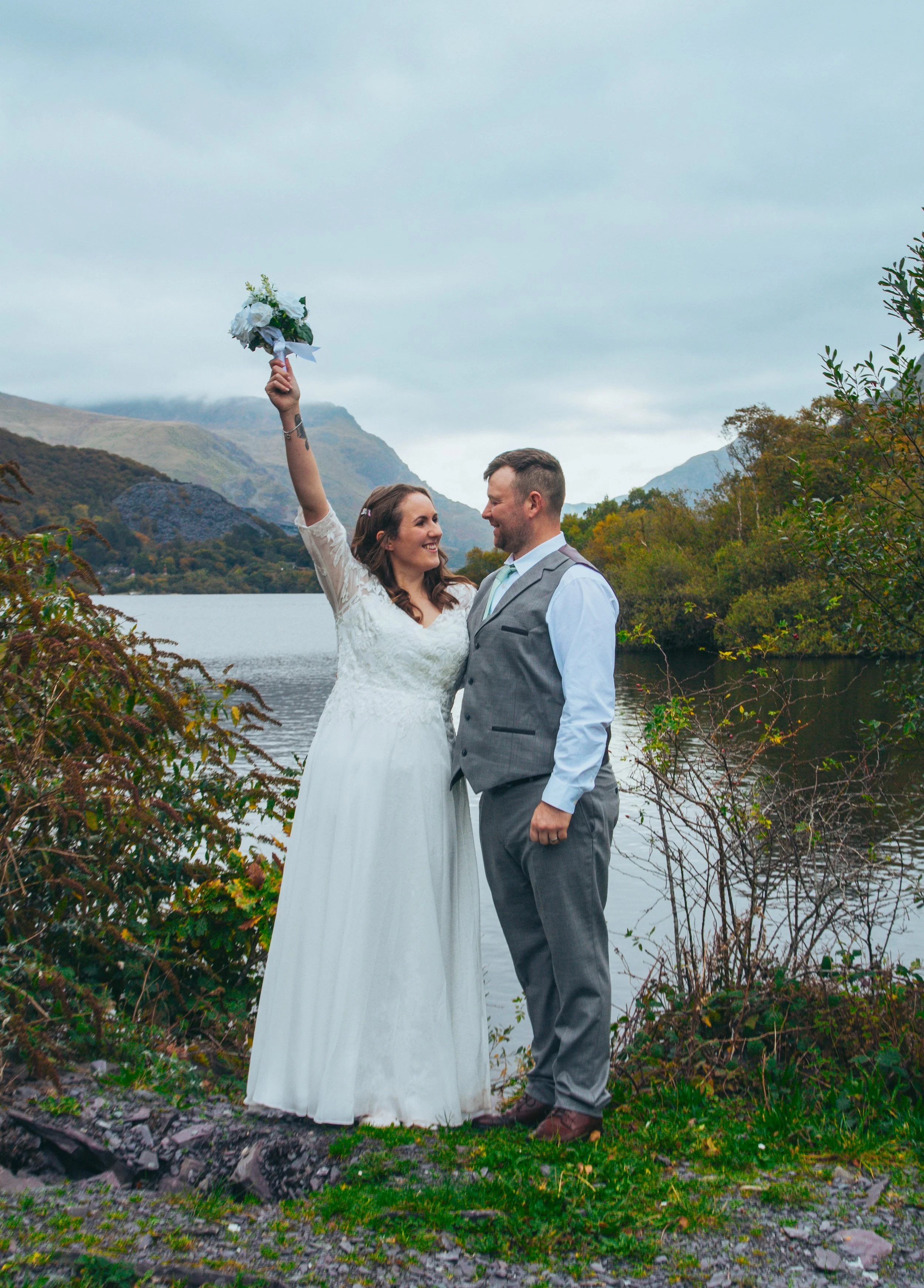 A bride raising her bouquet beside her groom on a lakeshore