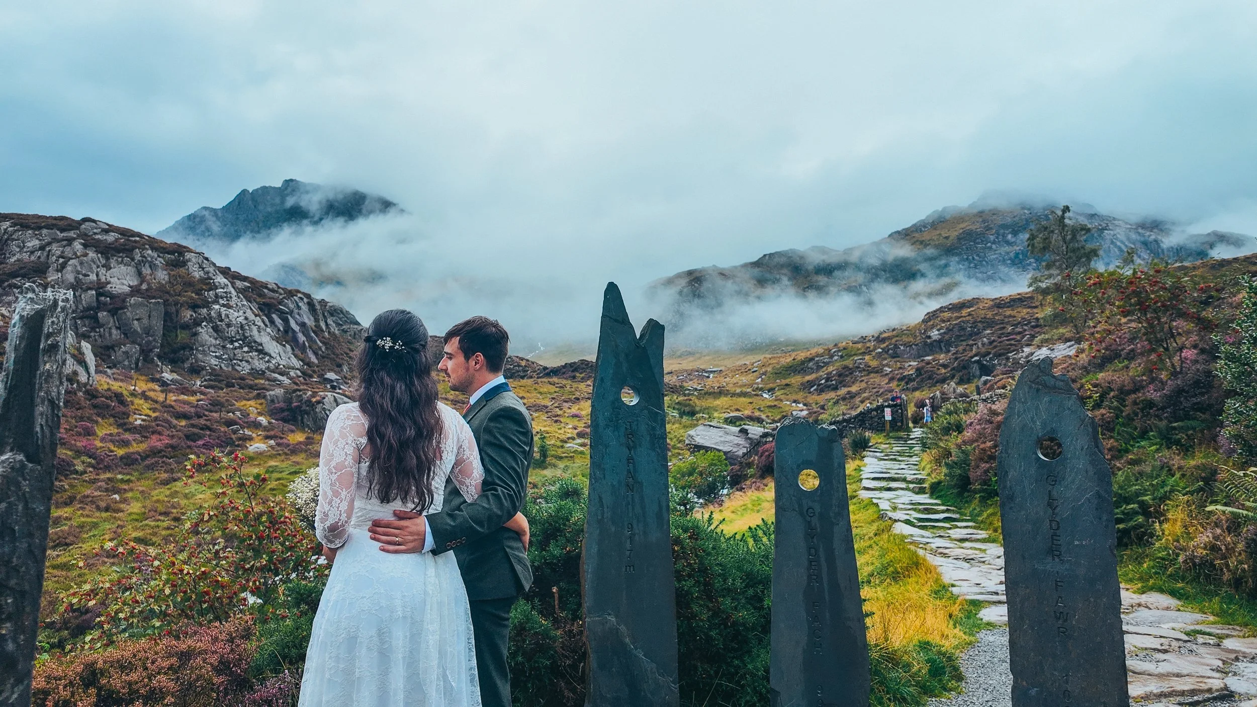 A couple embracing on a stone path between standing stones, with mist over the mountains