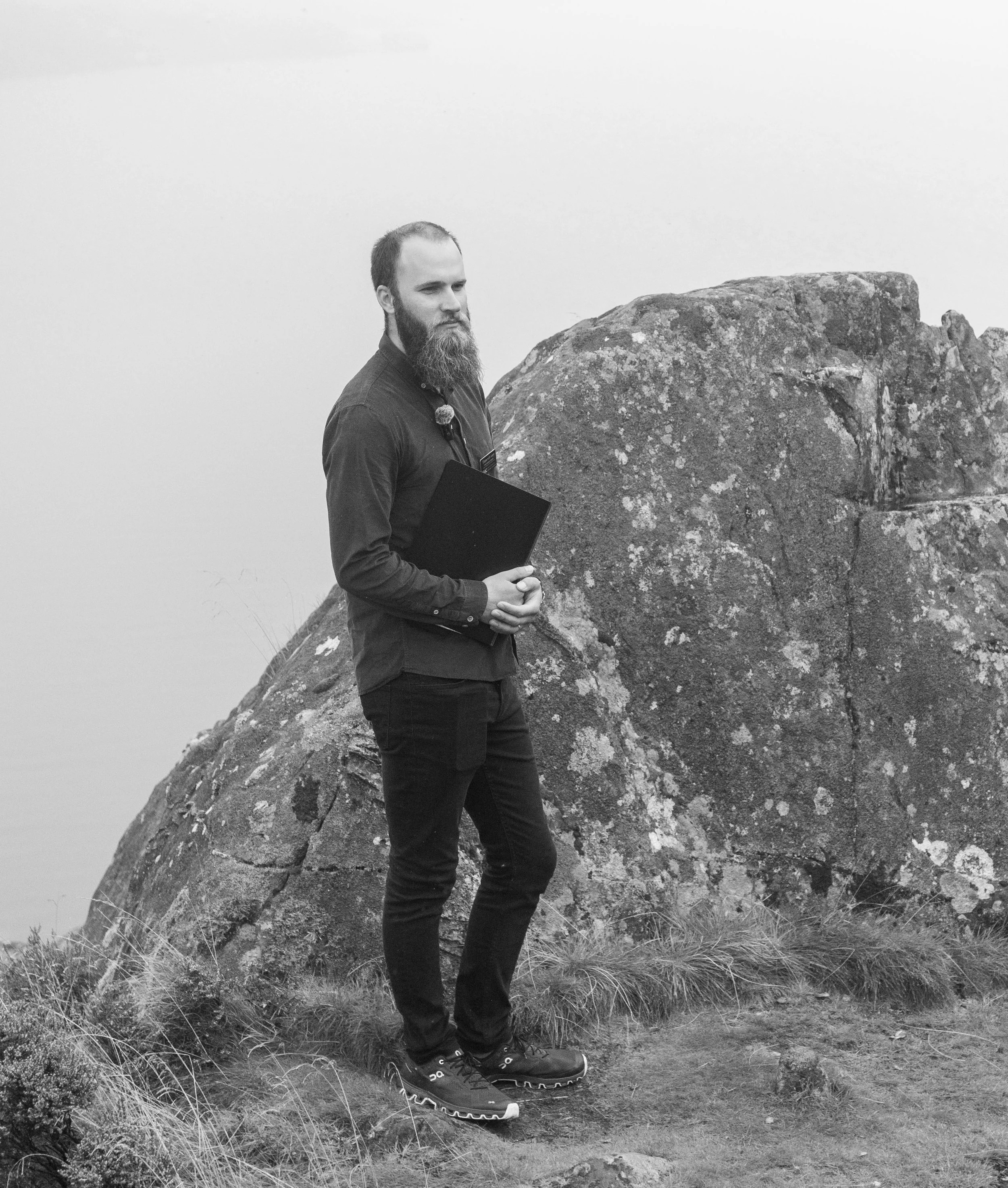Black and white photo of Leon holding a ceremony folder beside a rocky outcrop
