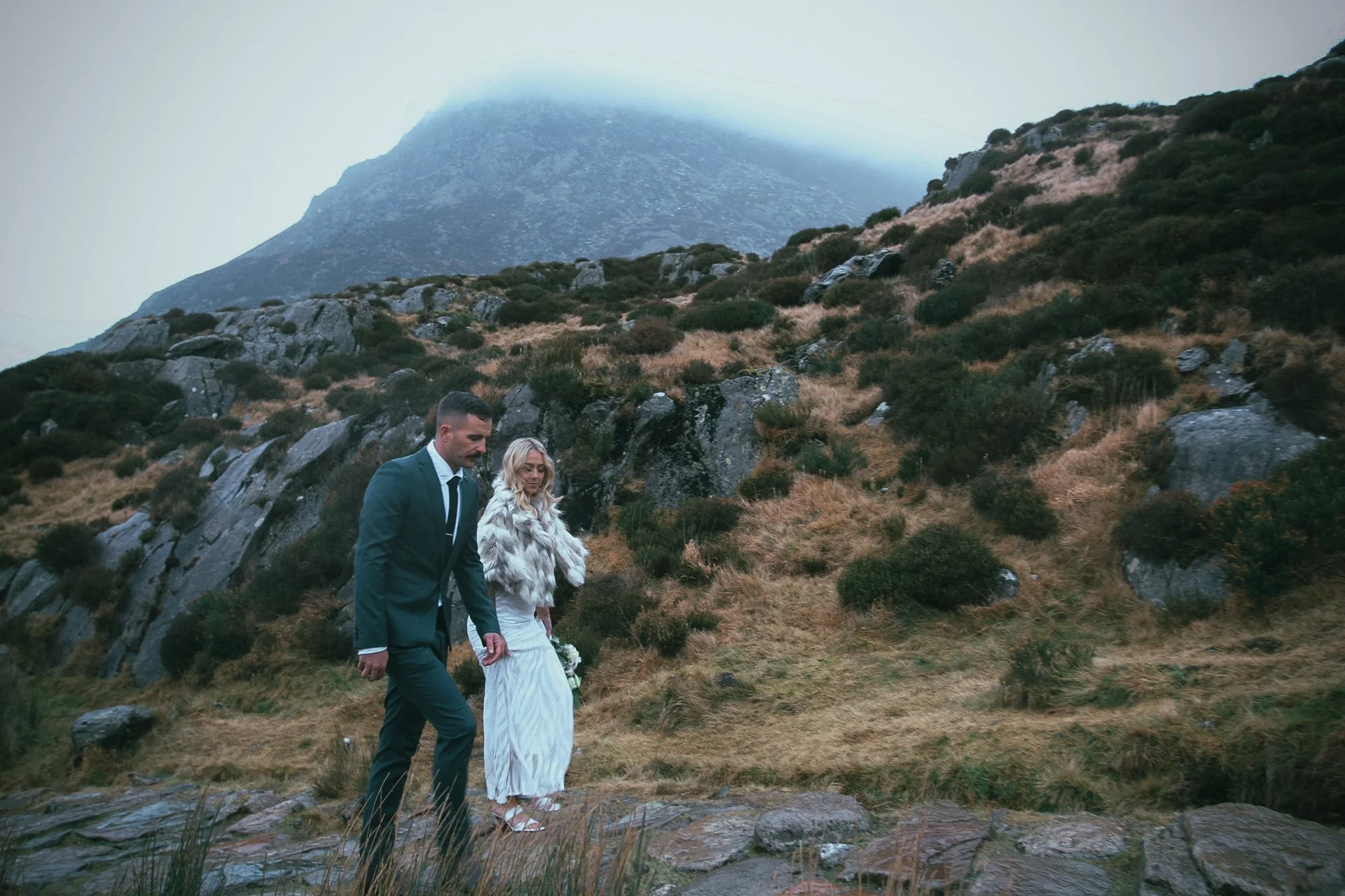 A couple standing on a rocky hillside beneath a misty mountain