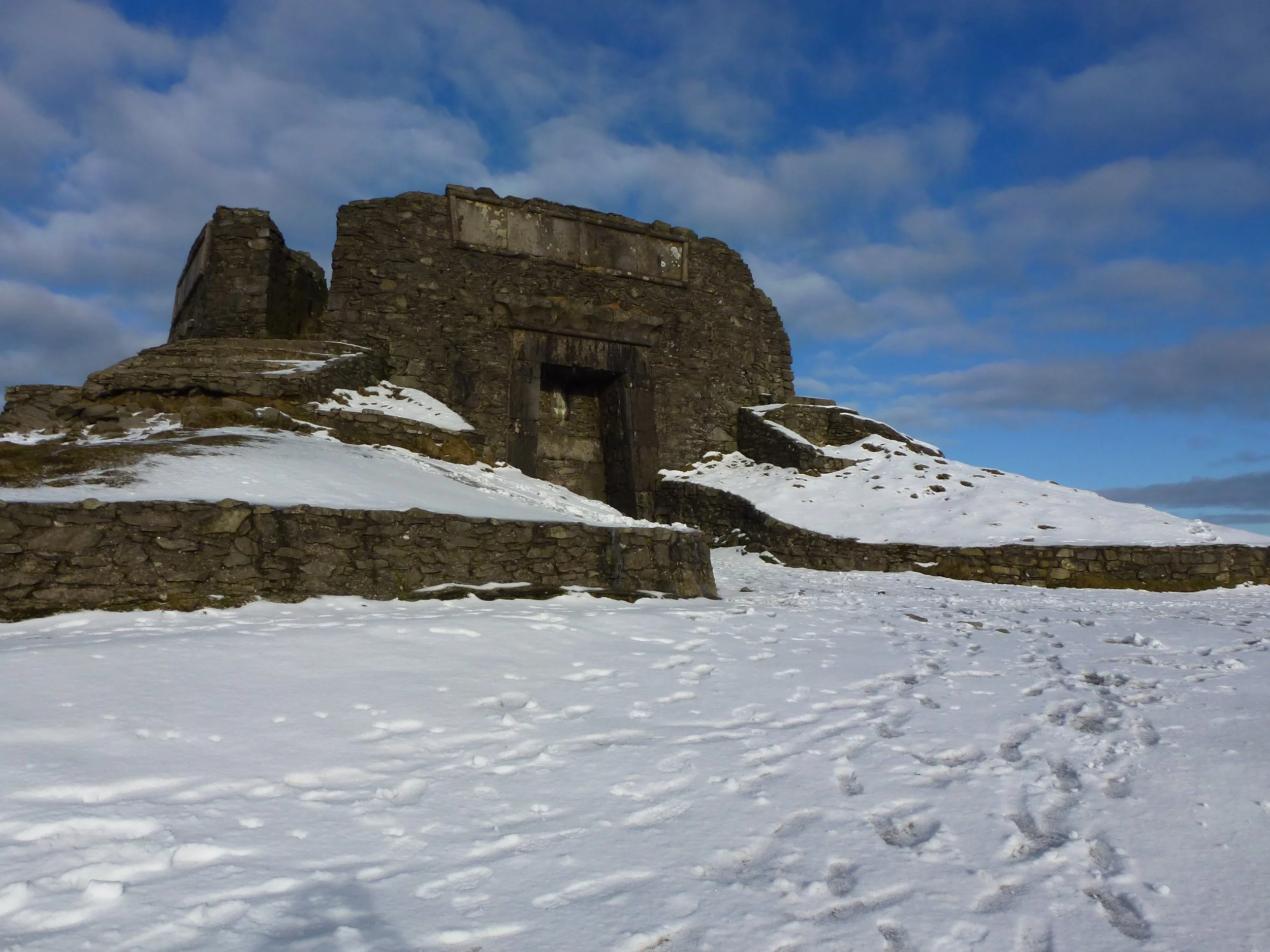 A ruined stone tower on a snow-covered hilltop beneath a blue sky with scattered clouds