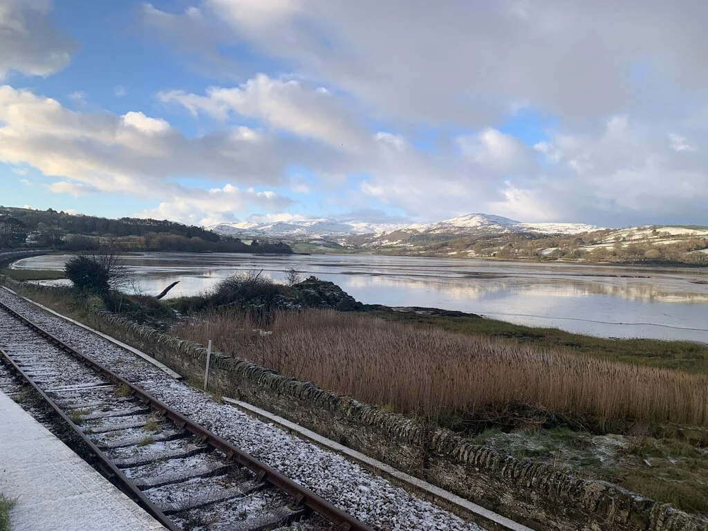 Snow-covered hills seen across a wide estuary from beside a railway line, under broken cloud