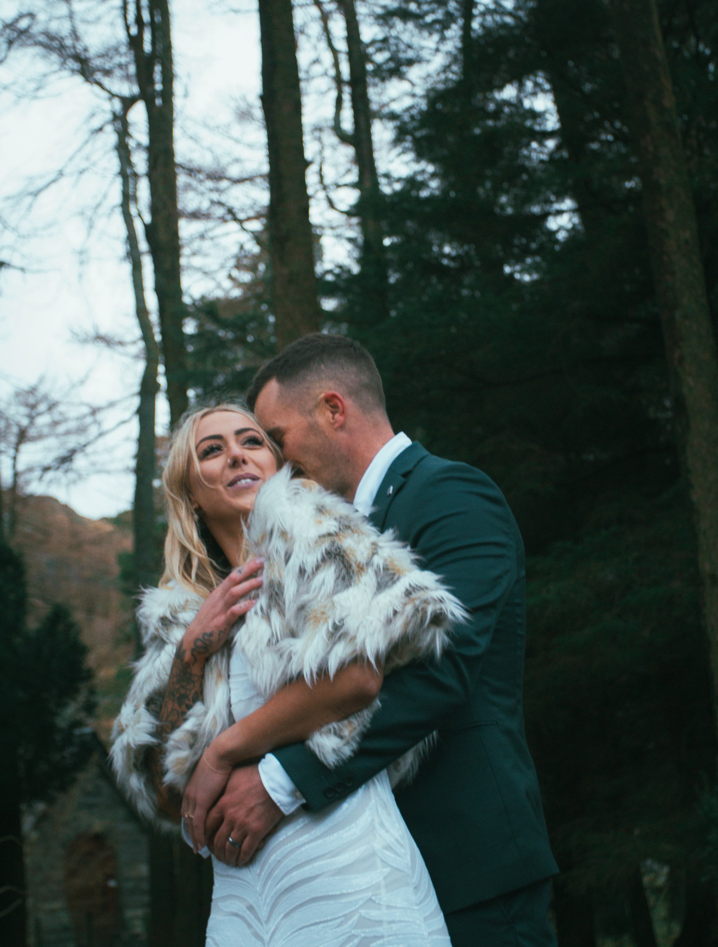 A groom embracing his bride from behind in a pine forest