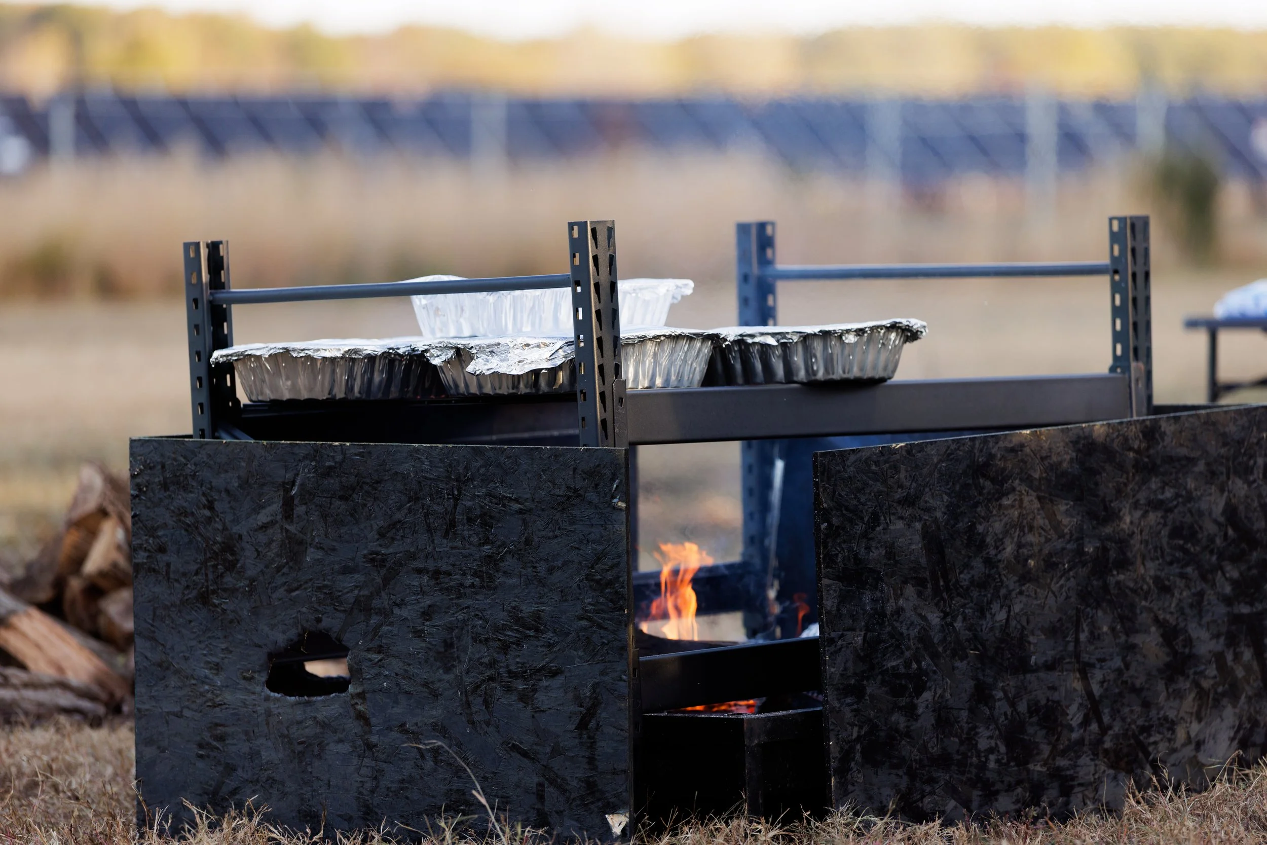 Outdoor cooking setup with a wood fire fire pit and a metal counter with aluminum trays and a glass bowl, situated on dry grass with a blurred background of a metal fence or structure.