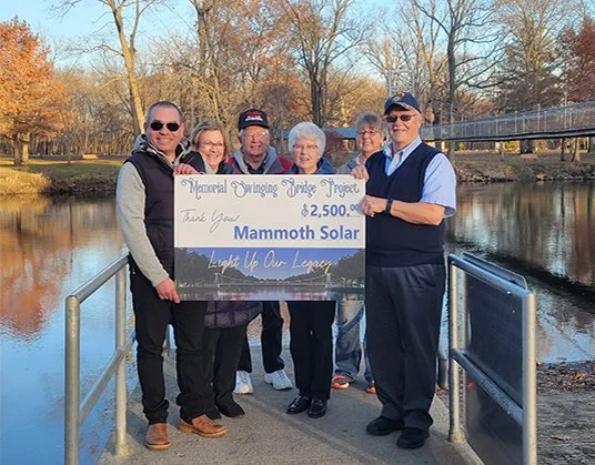 Group of six older adults standing on a small bridge by a pond, holding a large check for $2,500 from Mammoth Solar, outdoors with trees and a bridge in the background.