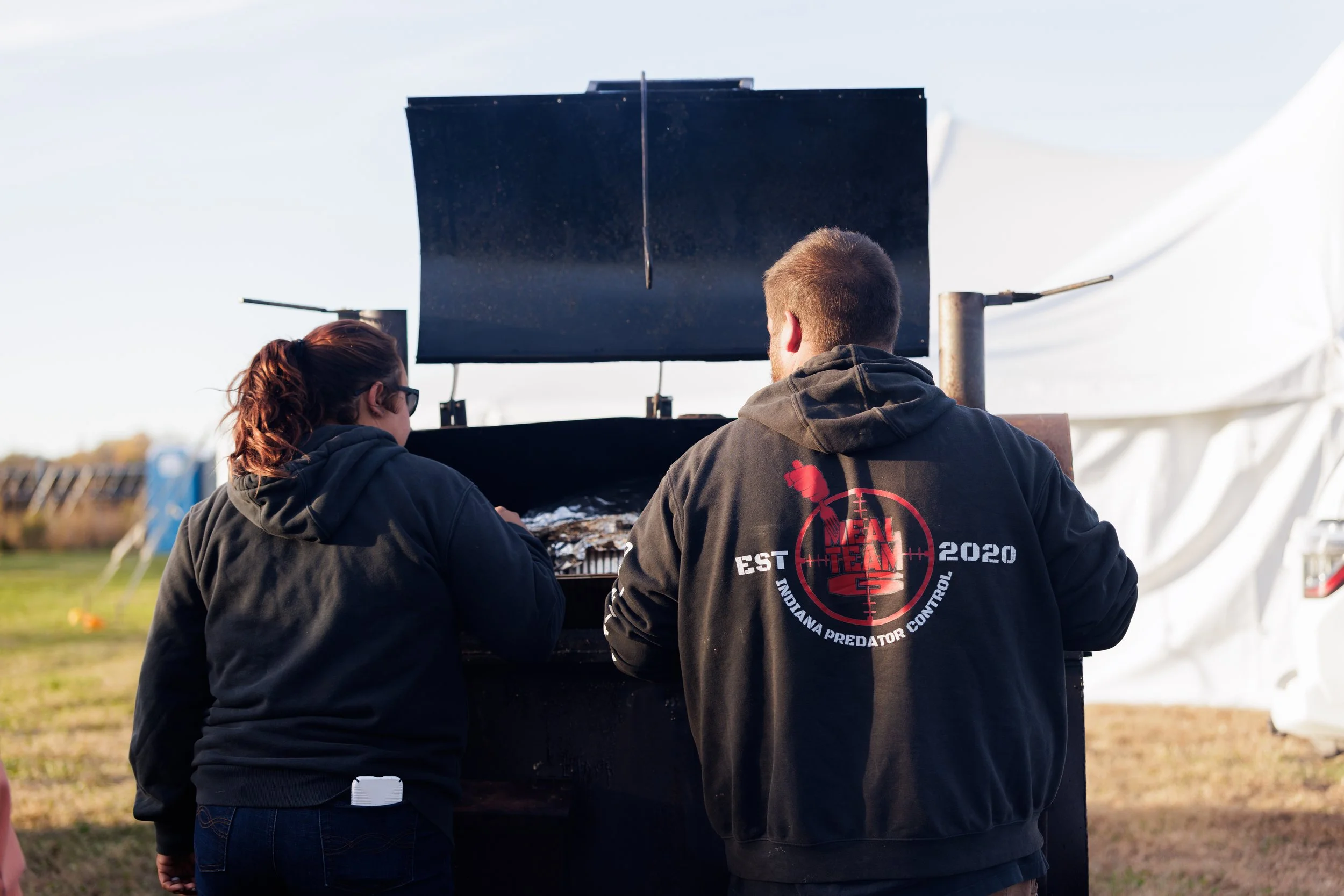 Two people grilling food outdoors during daytime, with a large flat grill and a white tent in the background.