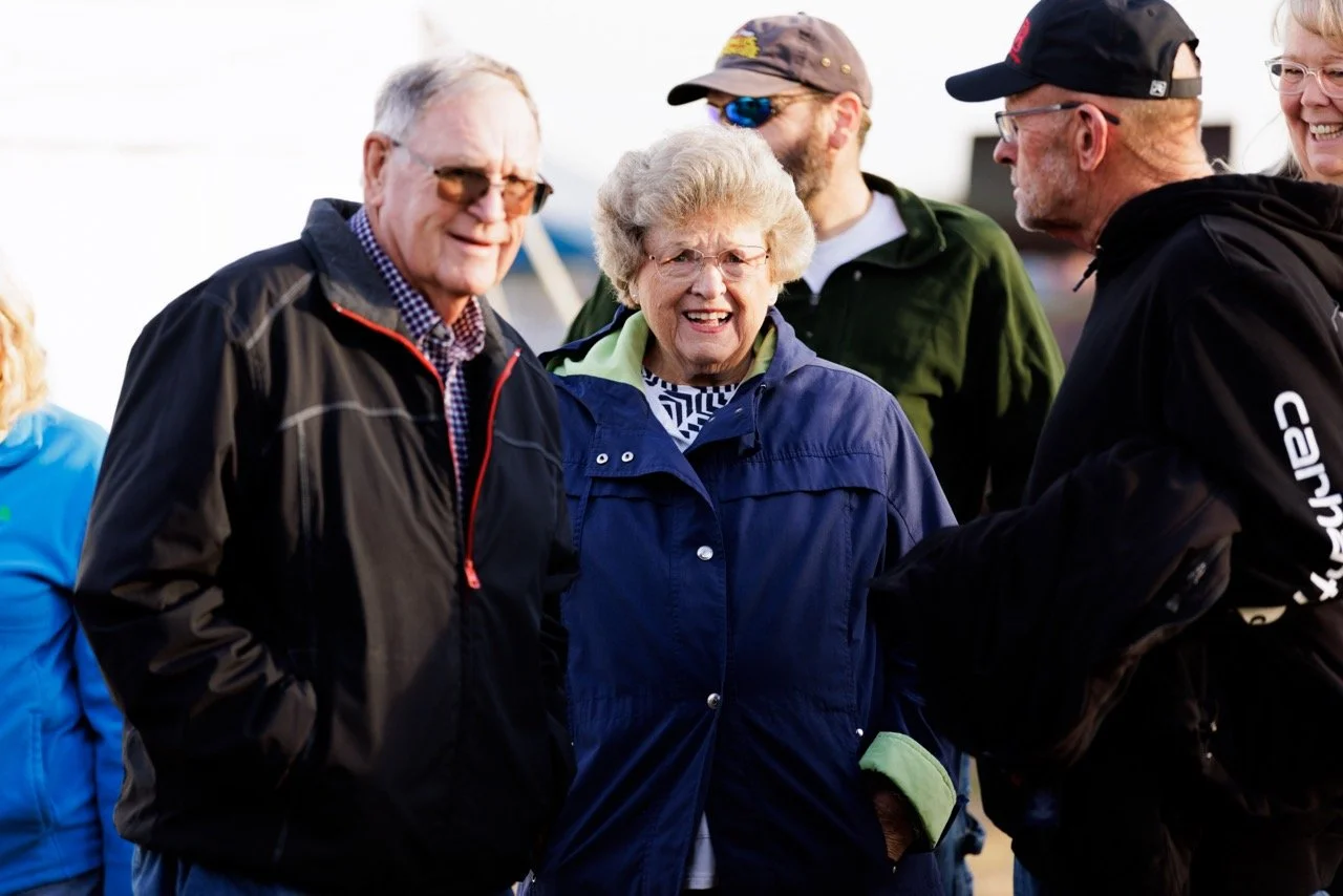 A group of smiling older adults outdoors, dressed in casual jackets and hats, engaging in a friendly conversation.