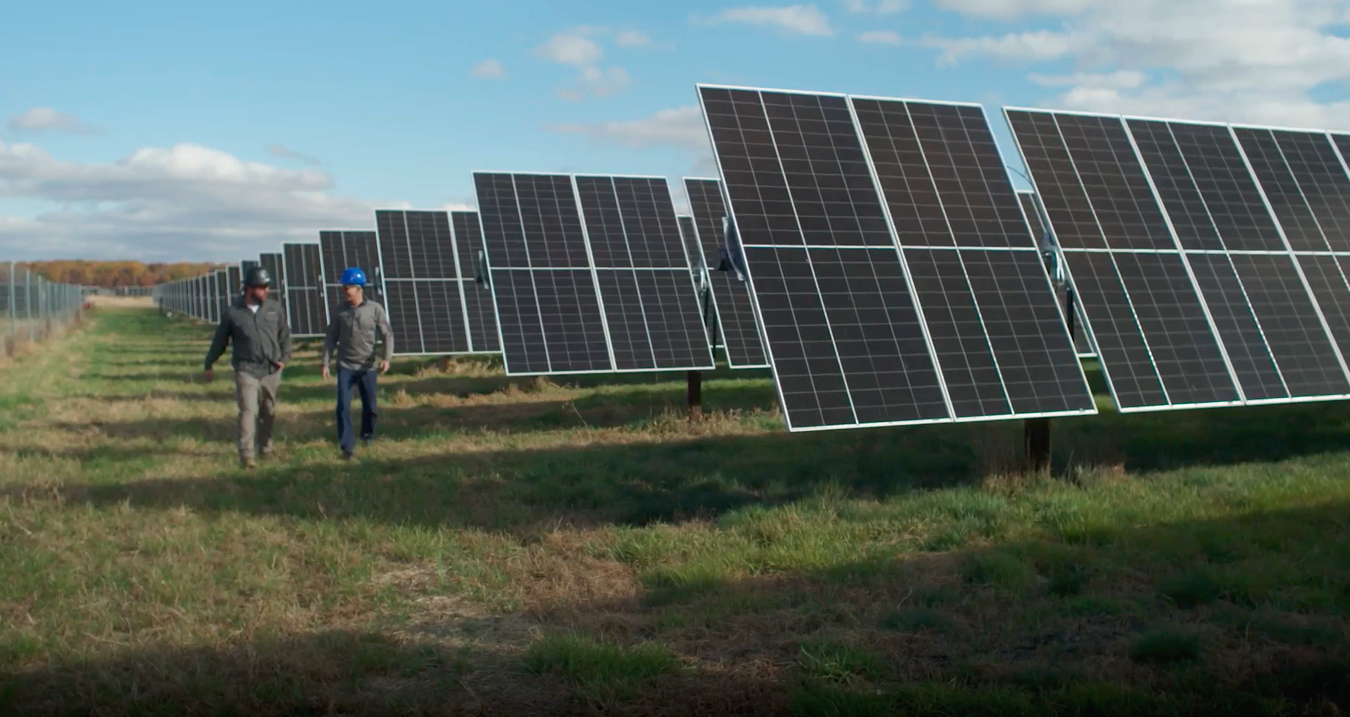 Two workers in hard hats walking through a solar farm with rows of solar panels on a partly cloudy day.