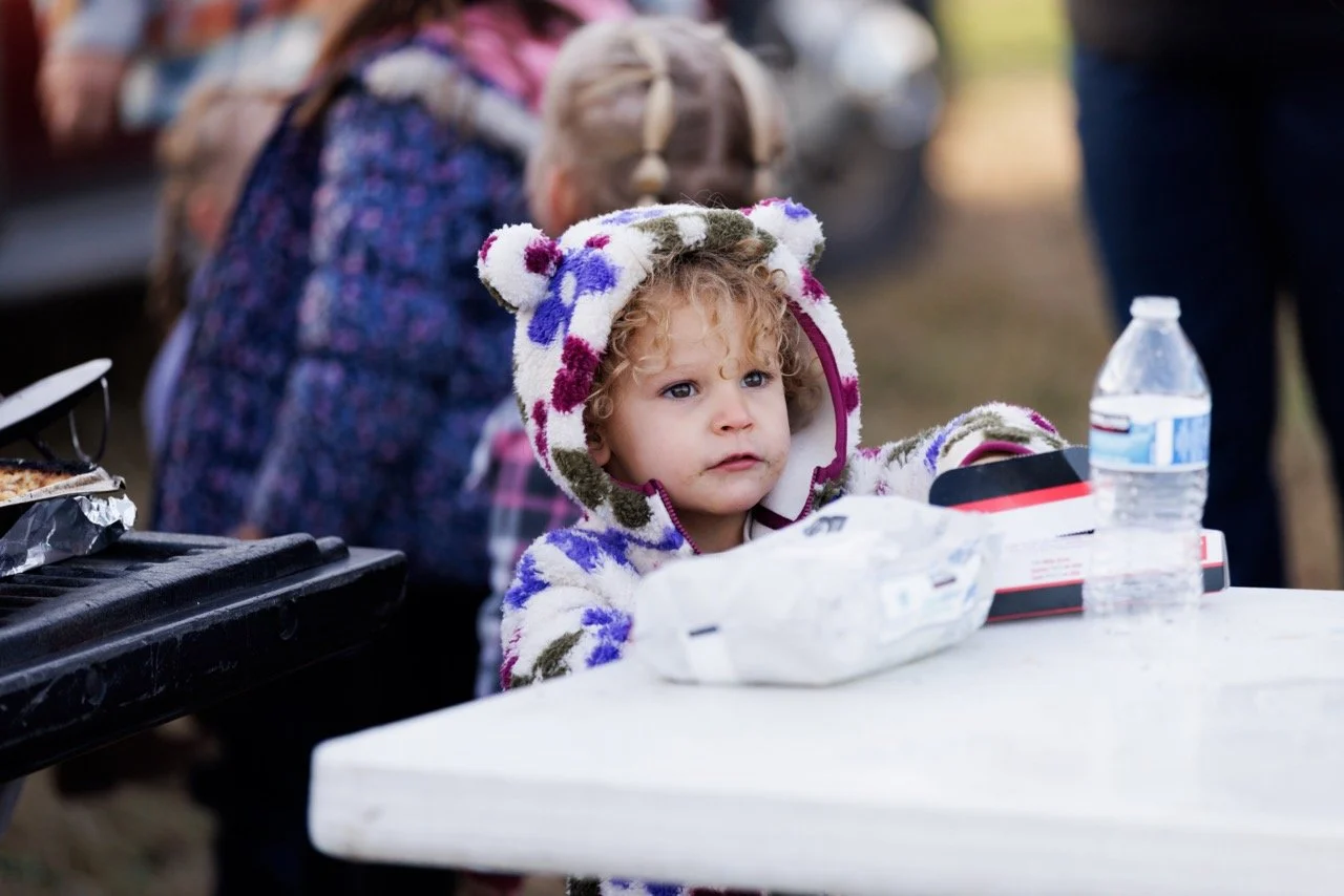 A young girl with curly hair wearing a hooded jacket with bear ears, sitting at a table outdoors with a water bottle, food container, and snack on the table, with people and cars in the background.