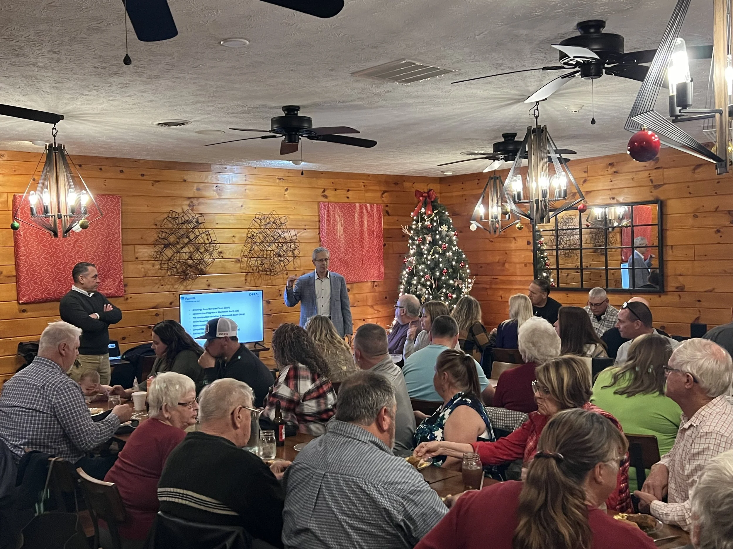 A man in a jacket gives a presentation to a seated audience in a room decorated for Christmas, with a Christmas tree and holiday decorations on the wooden walls.