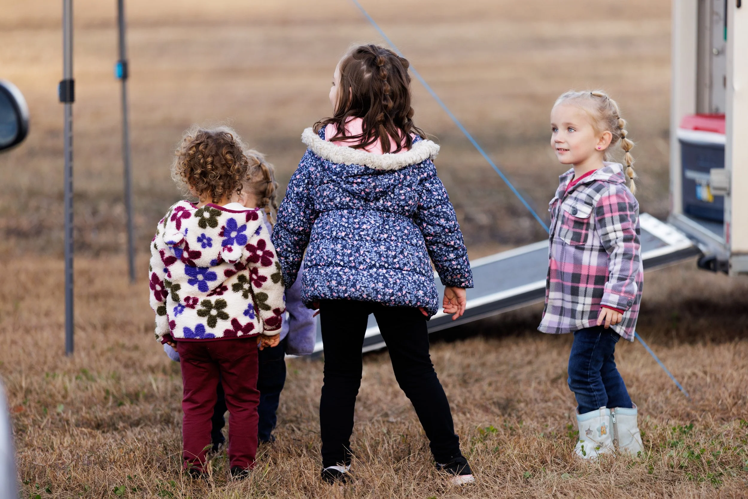 Four children standing outdoors on grass near a step ladder and a box, talking and facing each other.