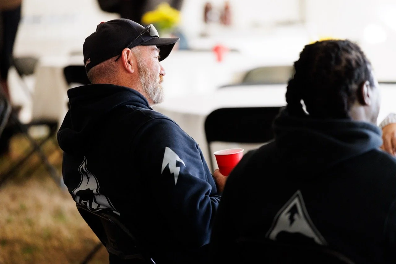 Two men sitting at a table, one with gray hair and beard wearing a black cap and hoodie, and the other with dark curly hair, both facing away. The man with the gray hair is holding a red cup.