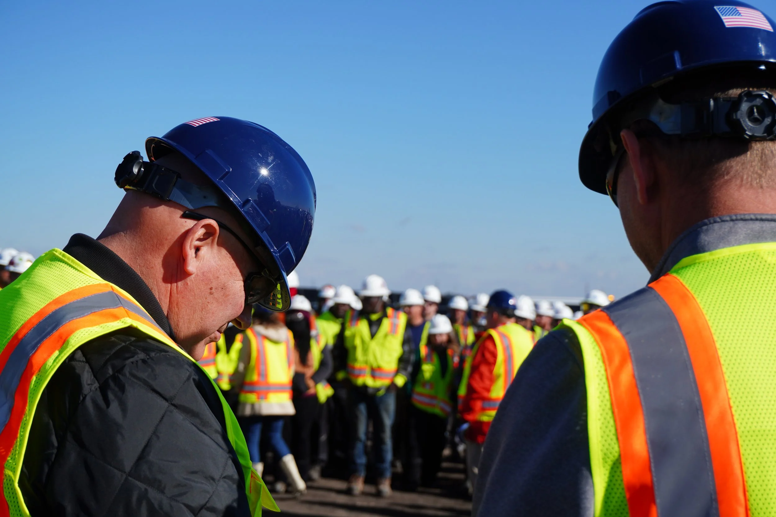 Two construction workers wearing blue helmets and high-visibility vests in foreground, with many other workers wearing similar gear in the background on a clear day.