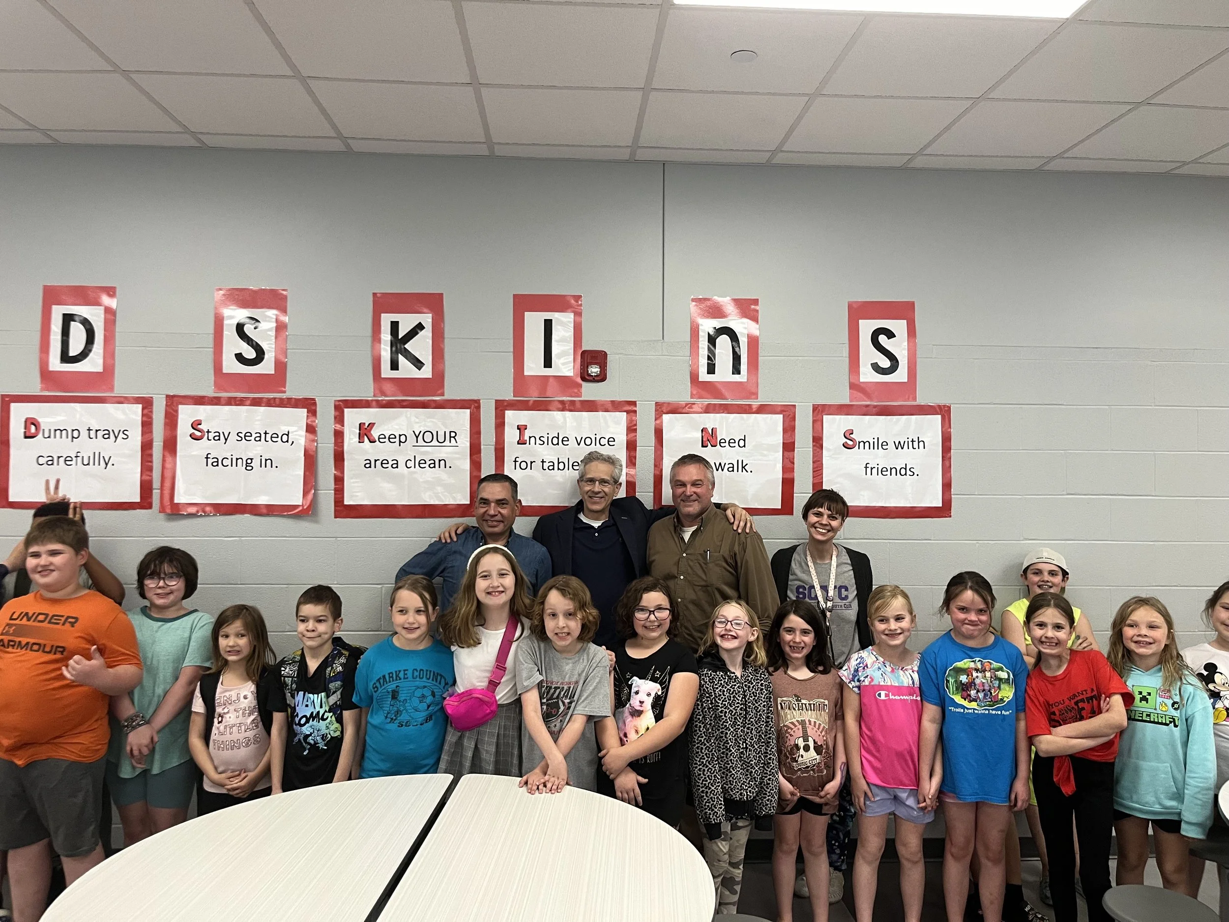 Group of children and three adults posing in front of a classroom wall decorated with letters and safety signs, with children smiling and making peace signs.