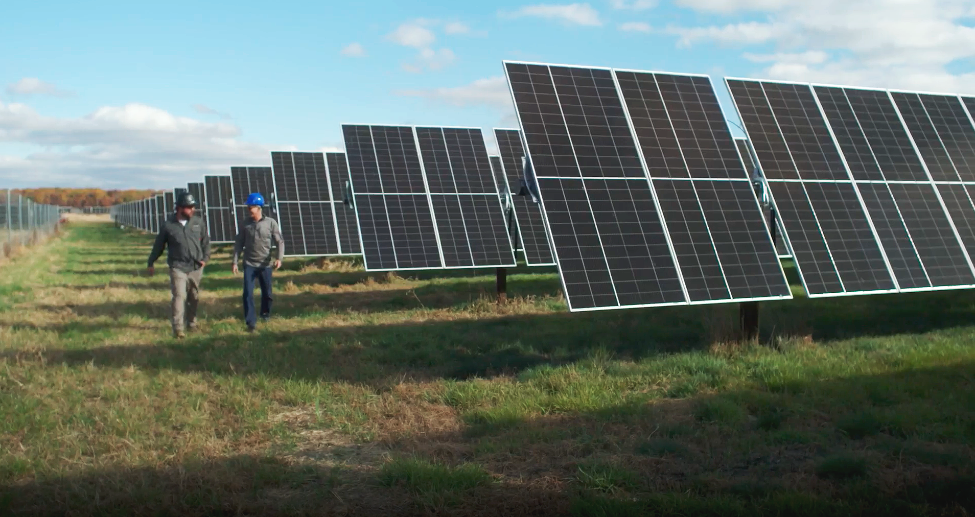 Two men walking and talking amidst a large field of solar panels on a partly cloudy day.