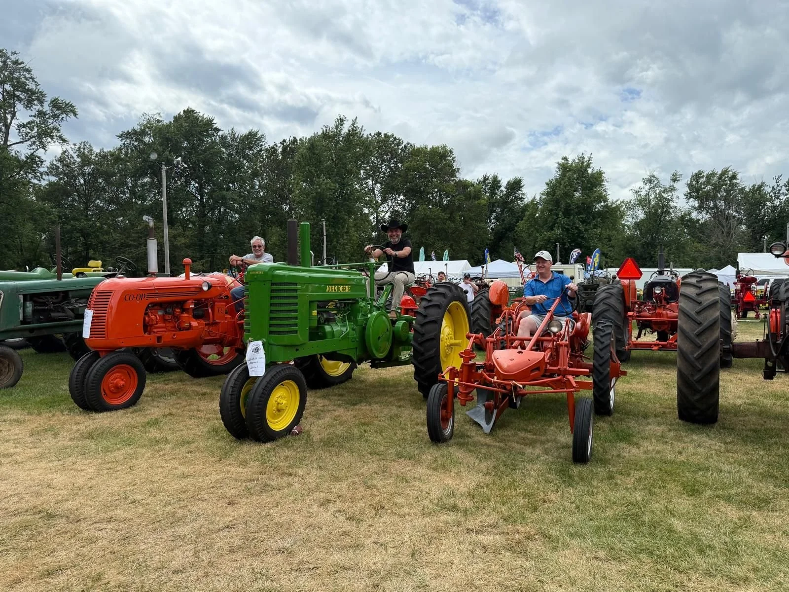 Three men sitting on vintage tractors at an outdoor event, with tents and other tractors in the background under a partly cloudy sky.