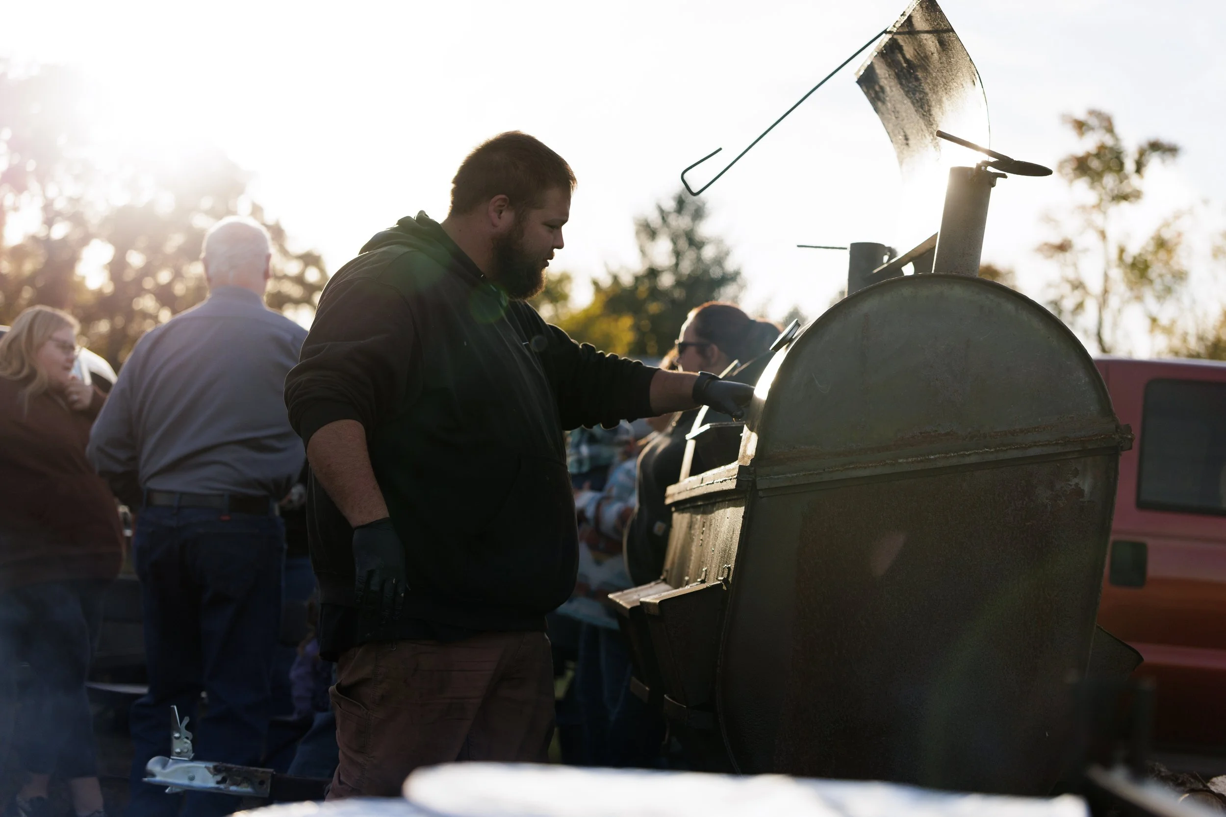 Man cooking on a large outdoor smoker grill at a gathering with several people, in the late afternoon sun, outdoors.