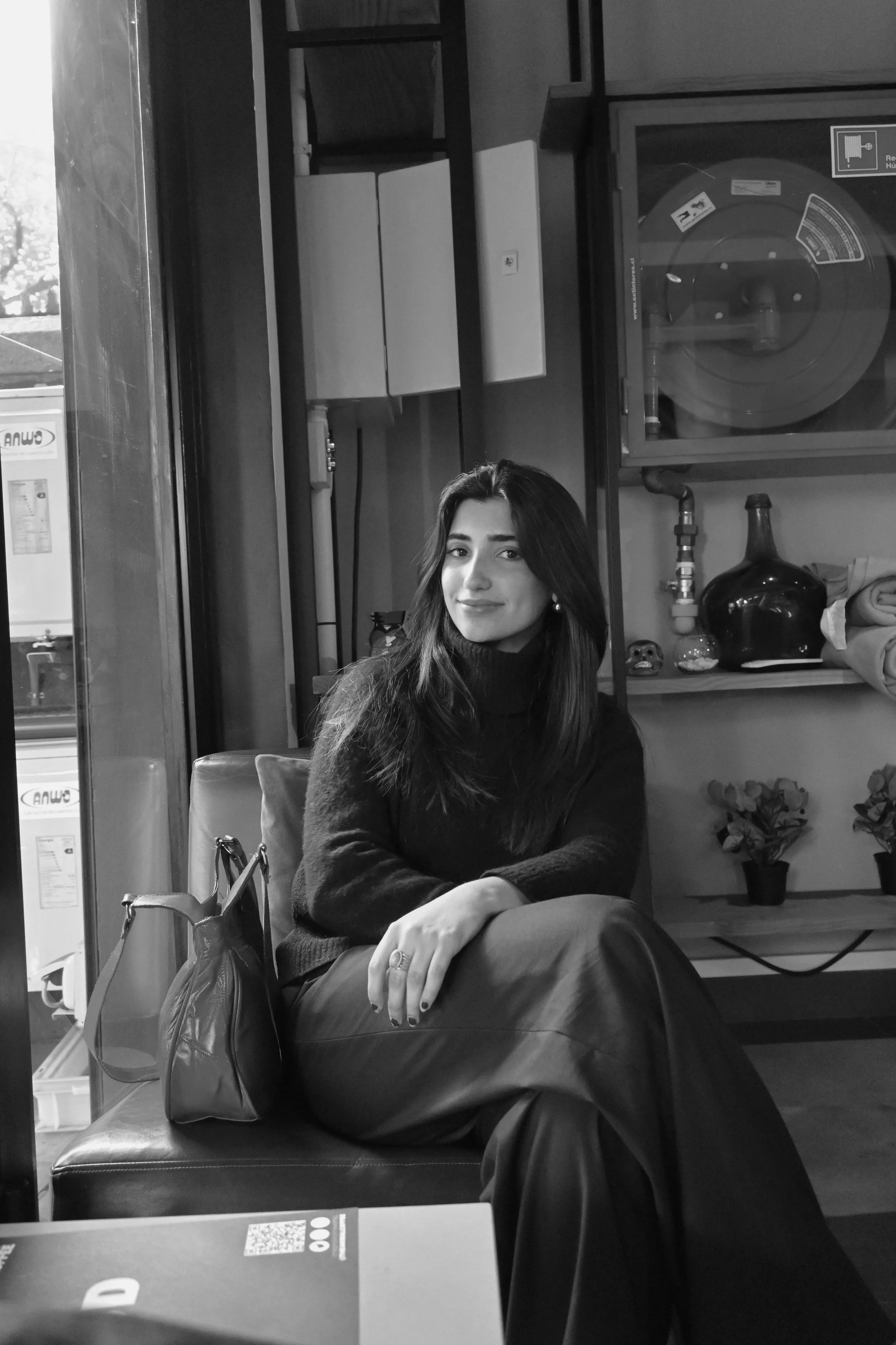 A woman with long dark hair, sitting on a booth in a cafe, wearing a black turtleneck sweater and dark wide-leg pants, with a bag beside her, smiling slightly at the camera.